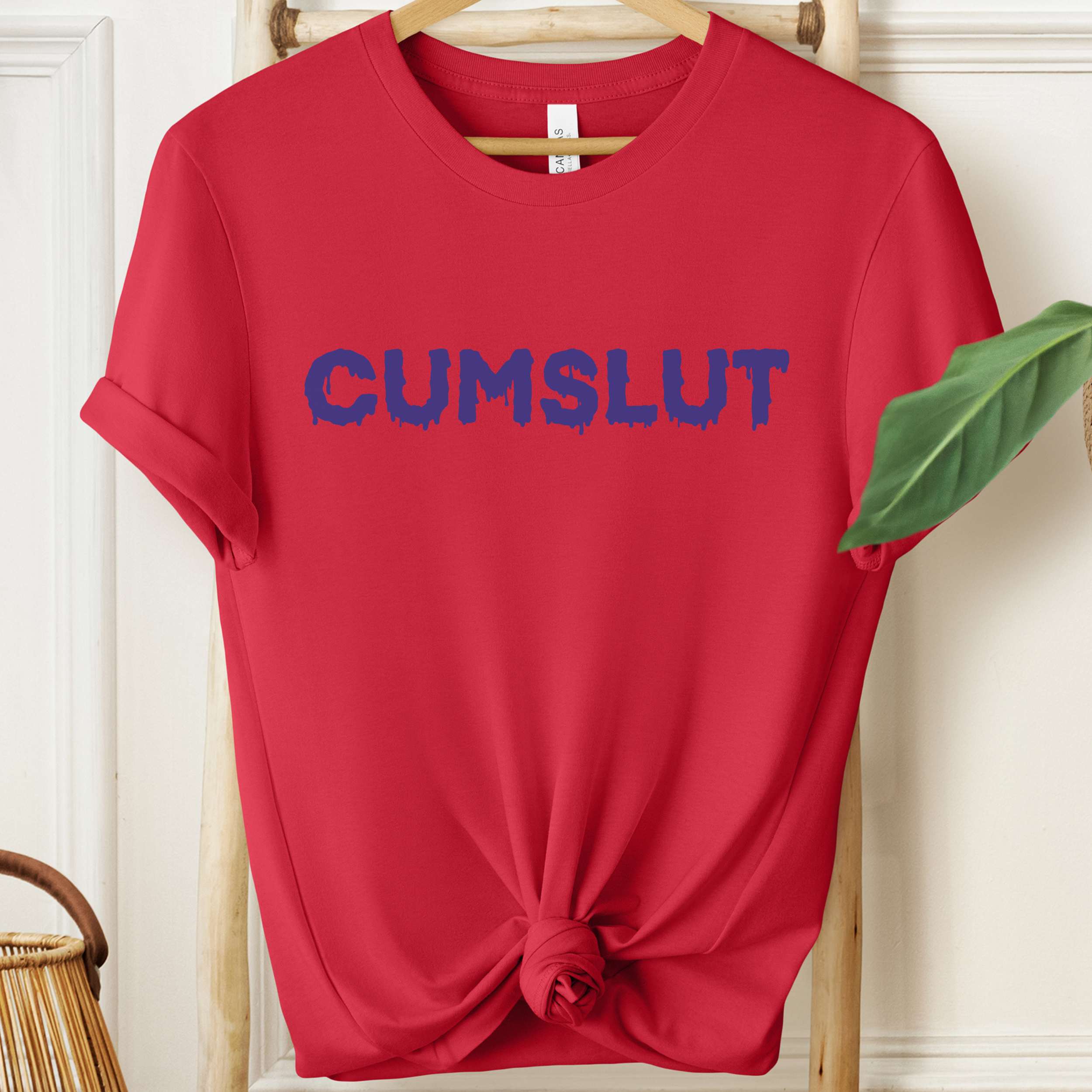 Red t-shirt with blue text 'CUMSLUT' on a hanger against a white wall.