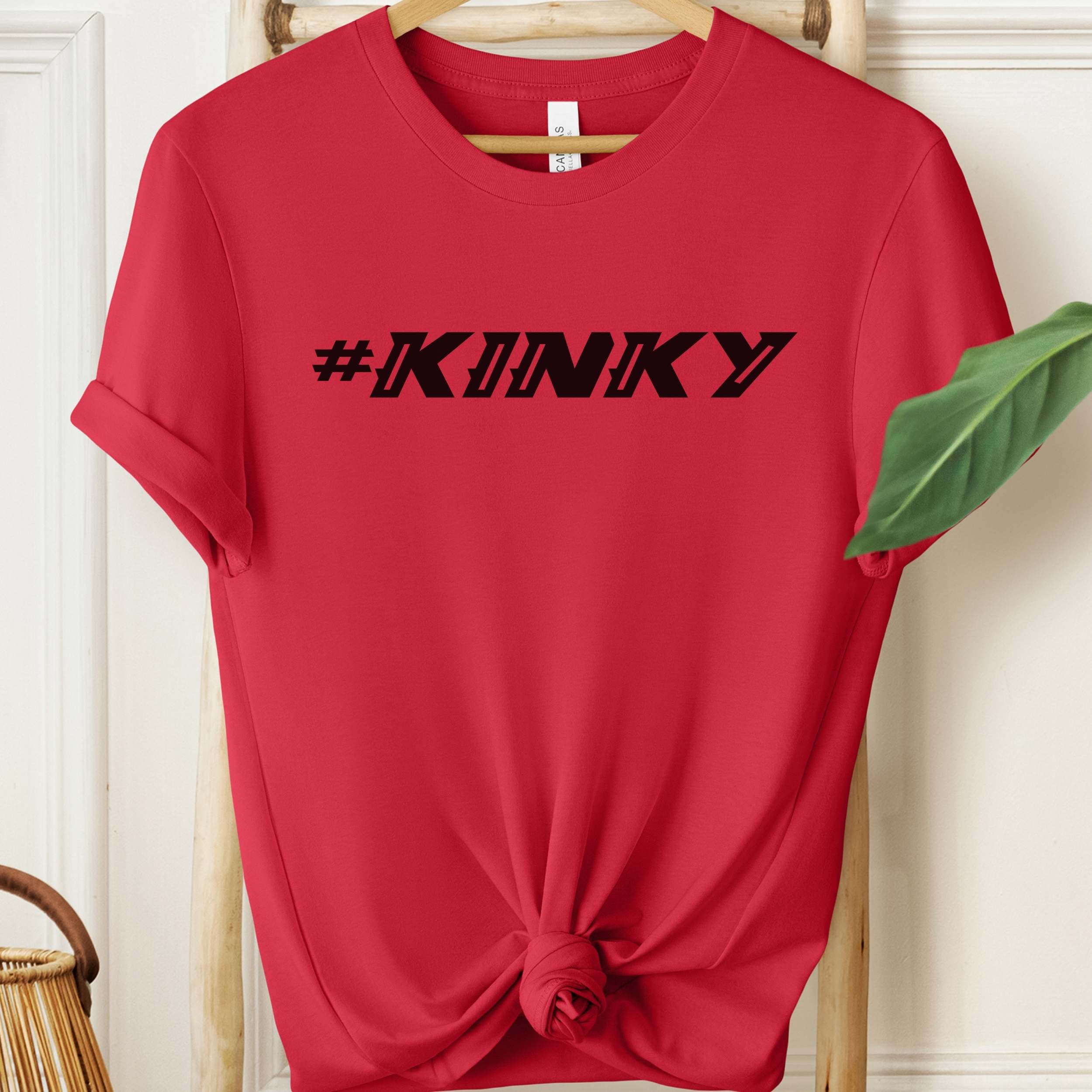 Red t-shirt with '#KINKY' text on a hanger against a white wall.