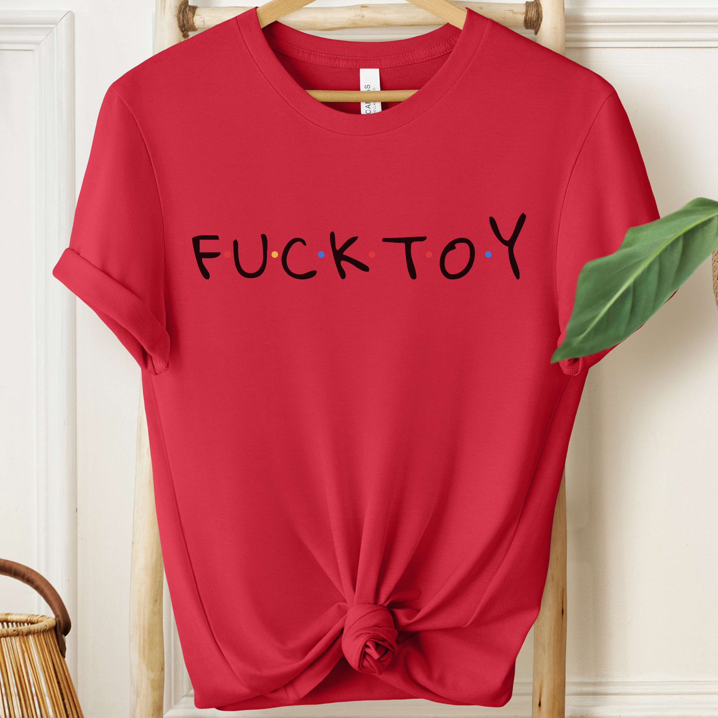 Red t-shirt with 'FUCK TOY' text on a hanger against a white wall.
