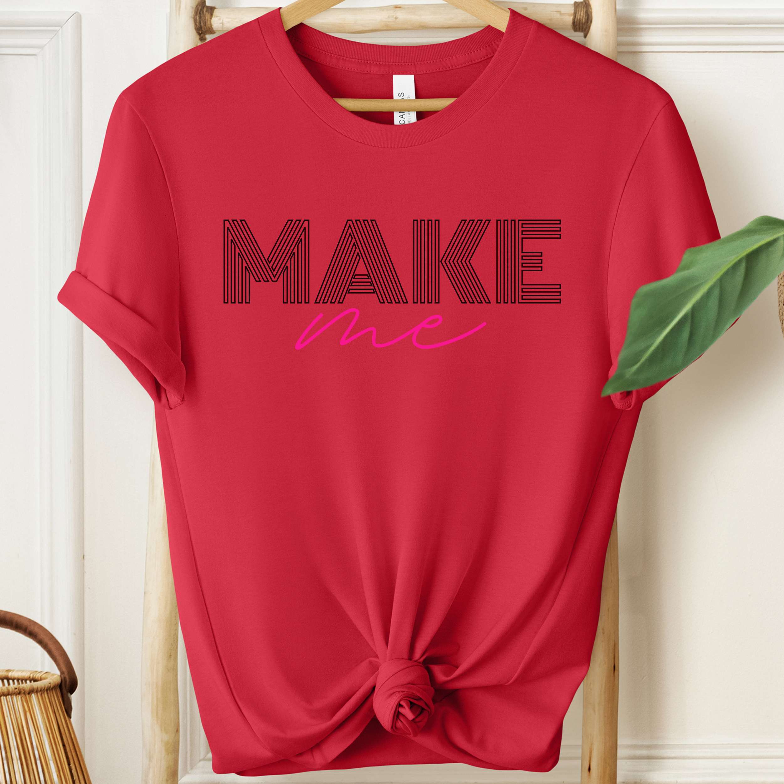 Red t-shirt with 'MAKE me' text on a hanger against a white wall.