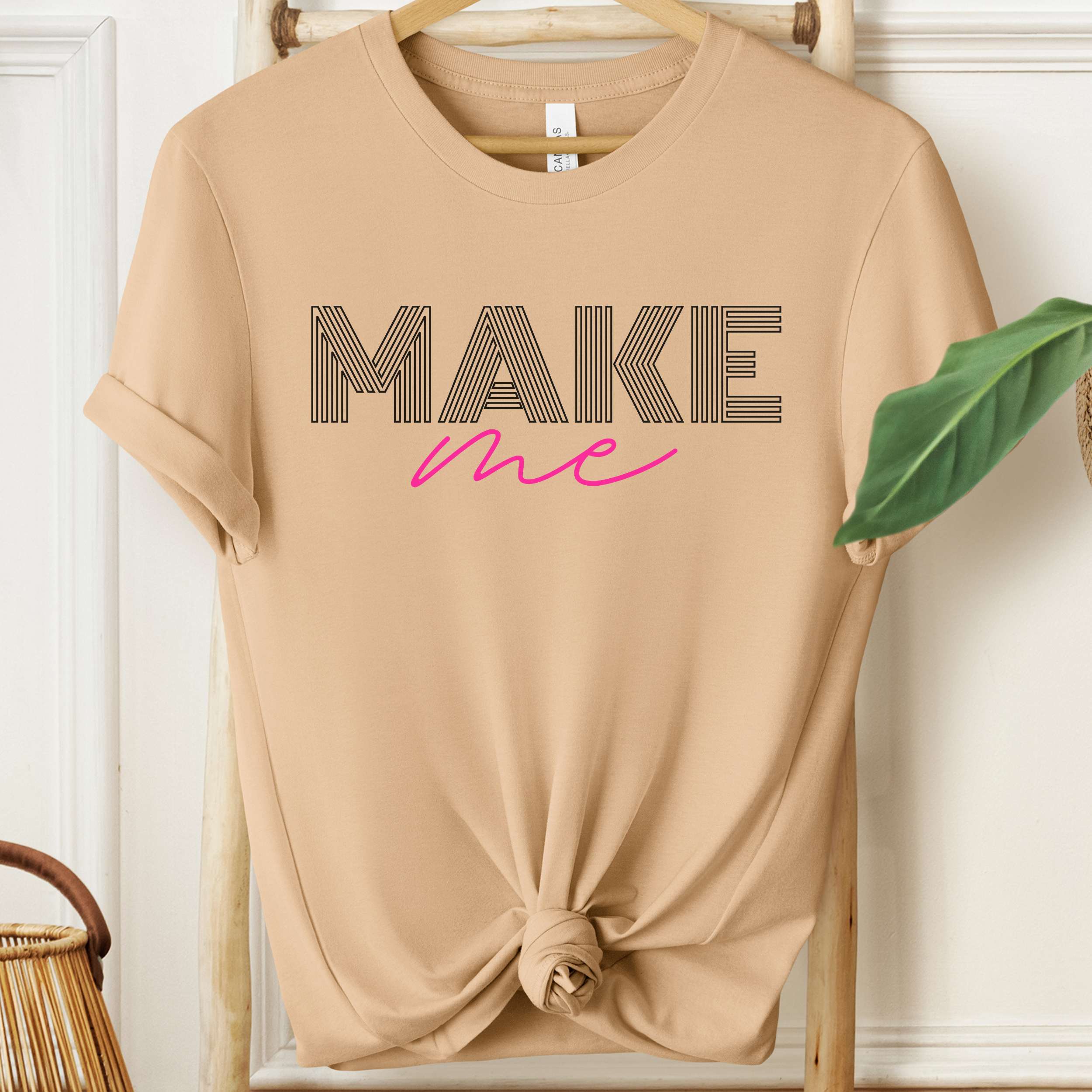 Beige t-shirt with 'MAKE me' text on a hanger against a white wall.