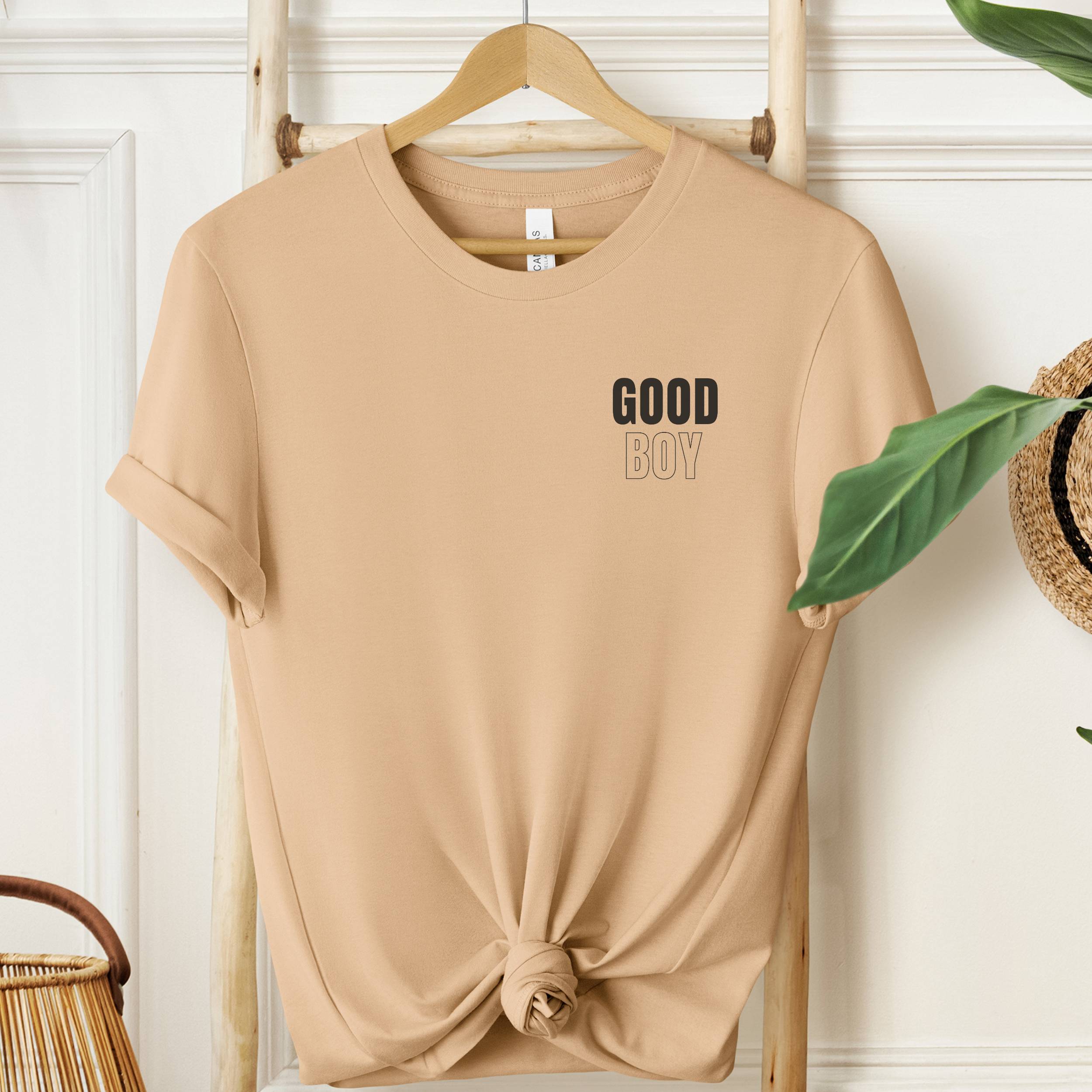 Beige t-shirt with 'GOOD BOY' text on a wooden hanger against a white wall.
