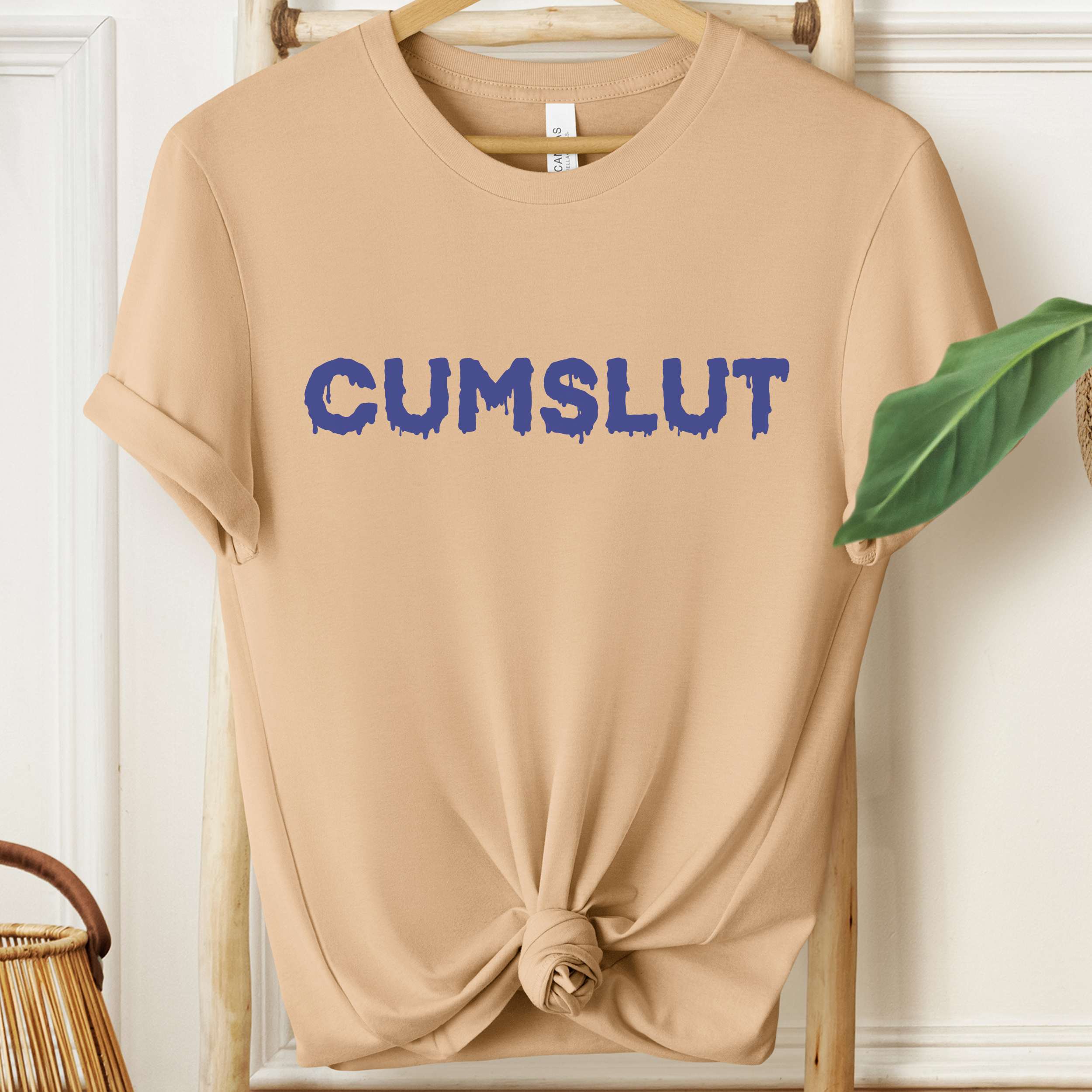 Beige t-shirt with blue text 'CUMSLUT' on a hanger against a white wall.