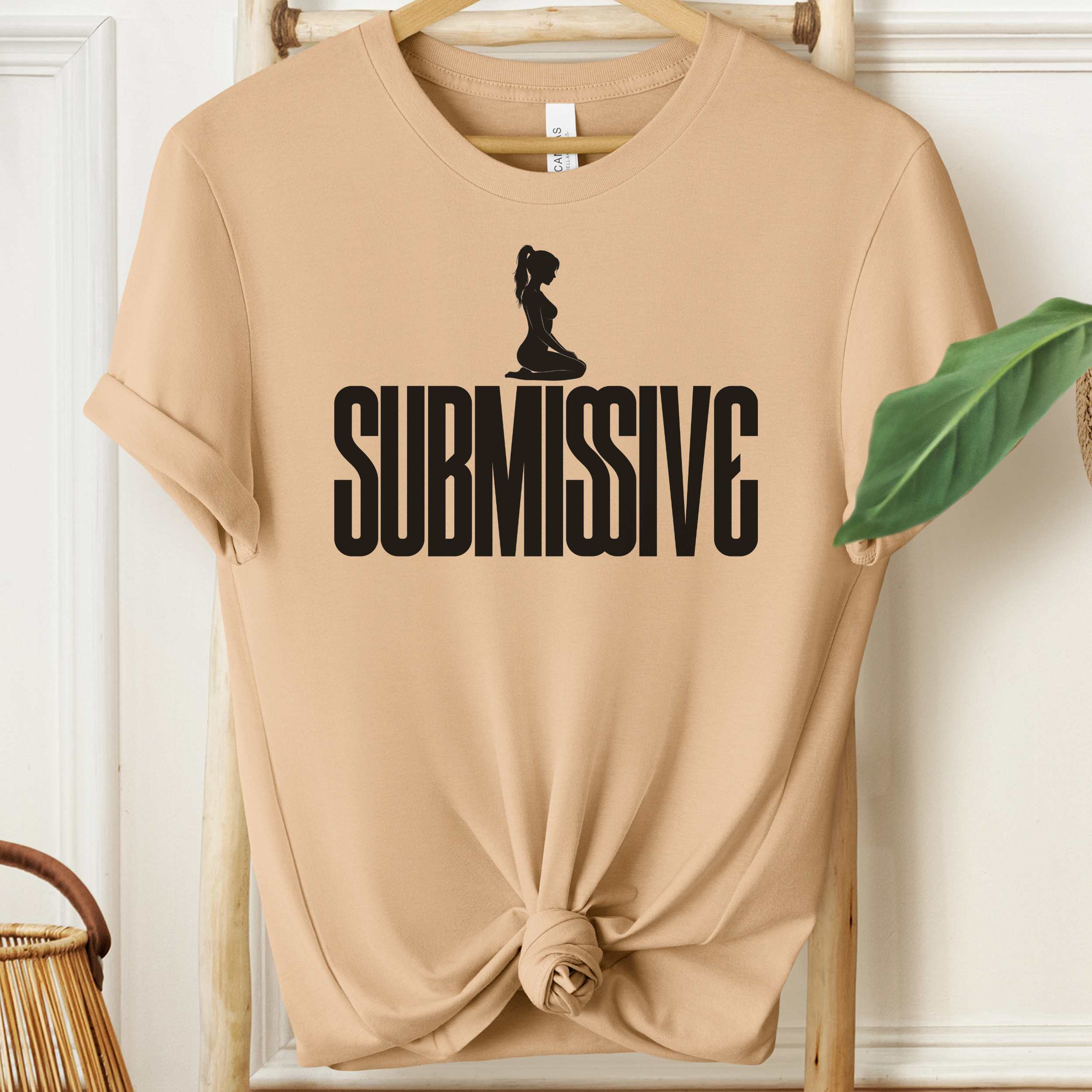 Beige t-shirt with 'SUBMISSIVE' graphic on a hanger against a white wall.