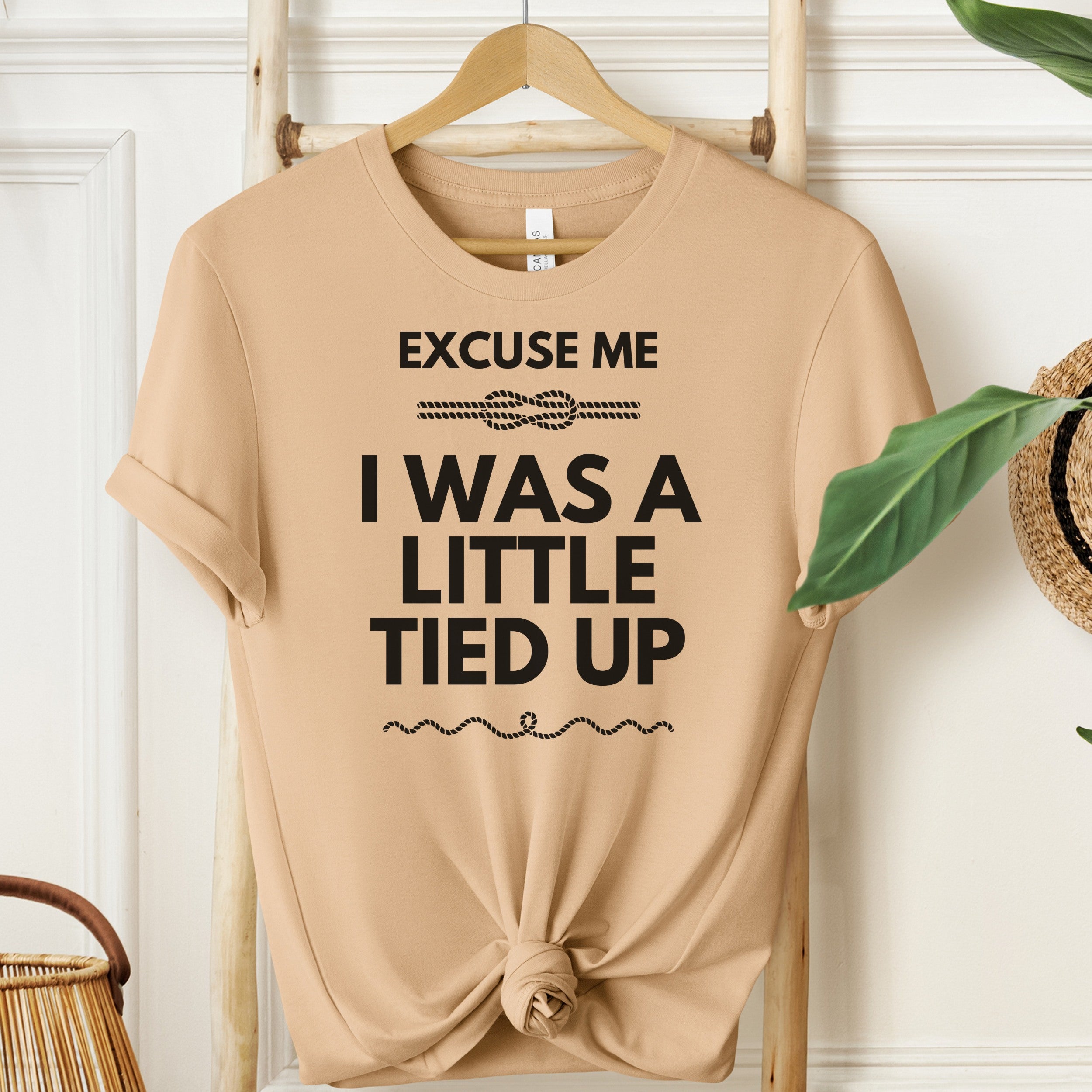 Beige t-shirt with humorous text on a wooden hanger against a white wall.
