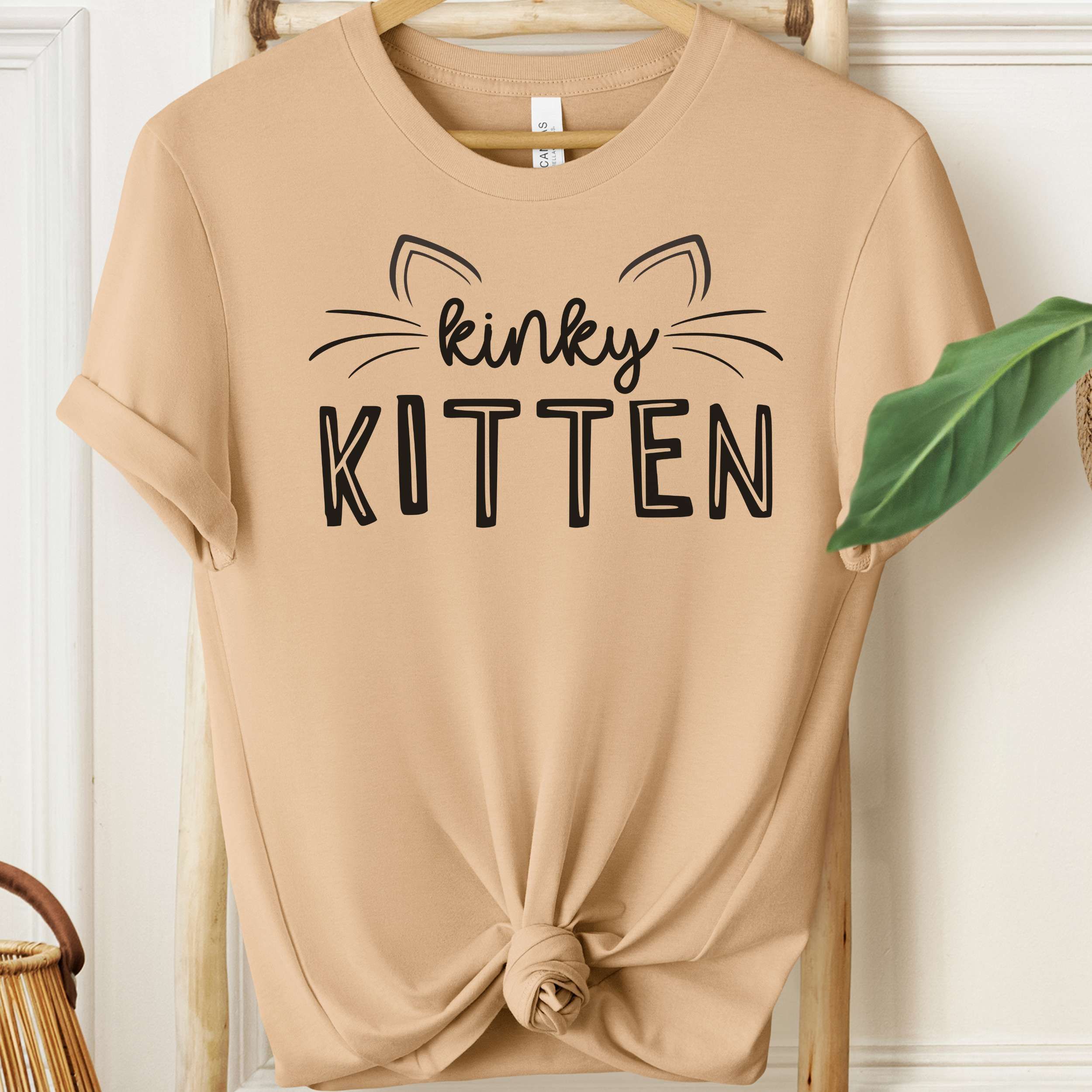 Beige t-shirt with 'kinky KITTEN' text on a wooden hanger.