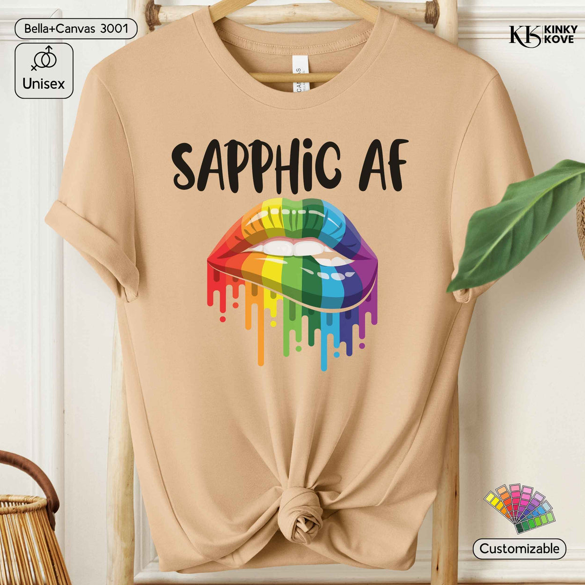 A beige short-sleeve t-shirt hanging on a wooden hanger, featuring a graphic of rainbow-colored dripping lips with the text 'SAPPHIC AF' in black above it. The shirt is styled with a knot at the bottom hem, set against a light wood panel background with decorative props.