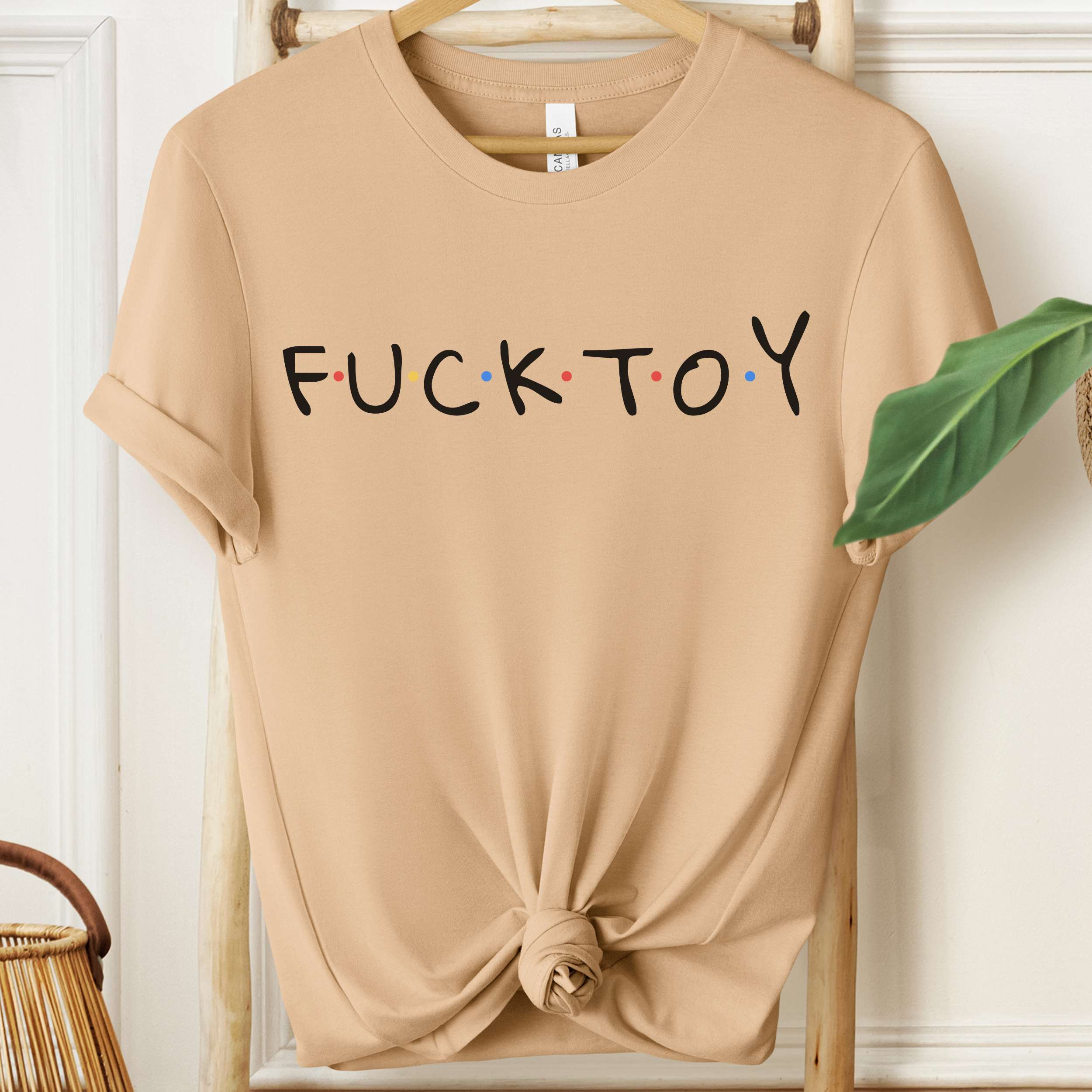 Beige t-shirt with 'FUCK TOY' text on a hanger against a white wall.