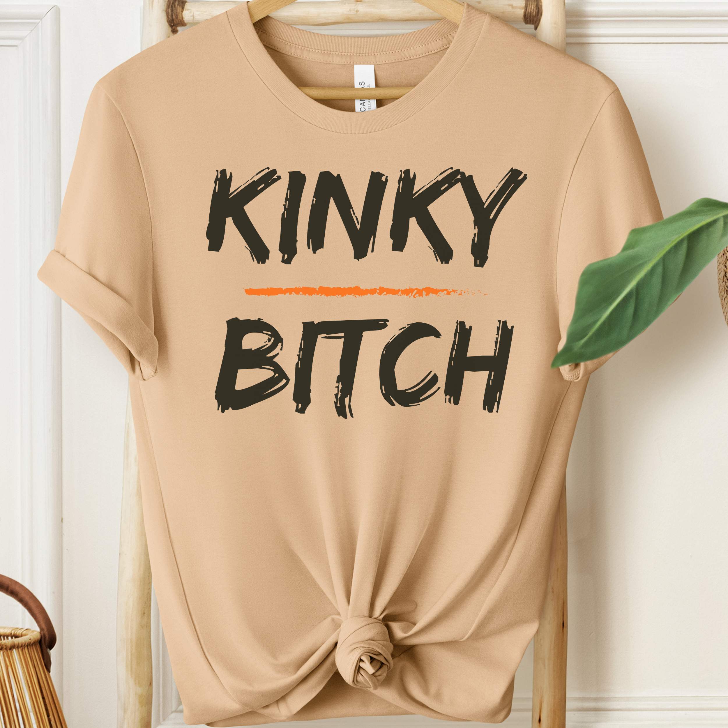 Beige t-shirt with 'KINKY BITCH' text on a wooden hanger.