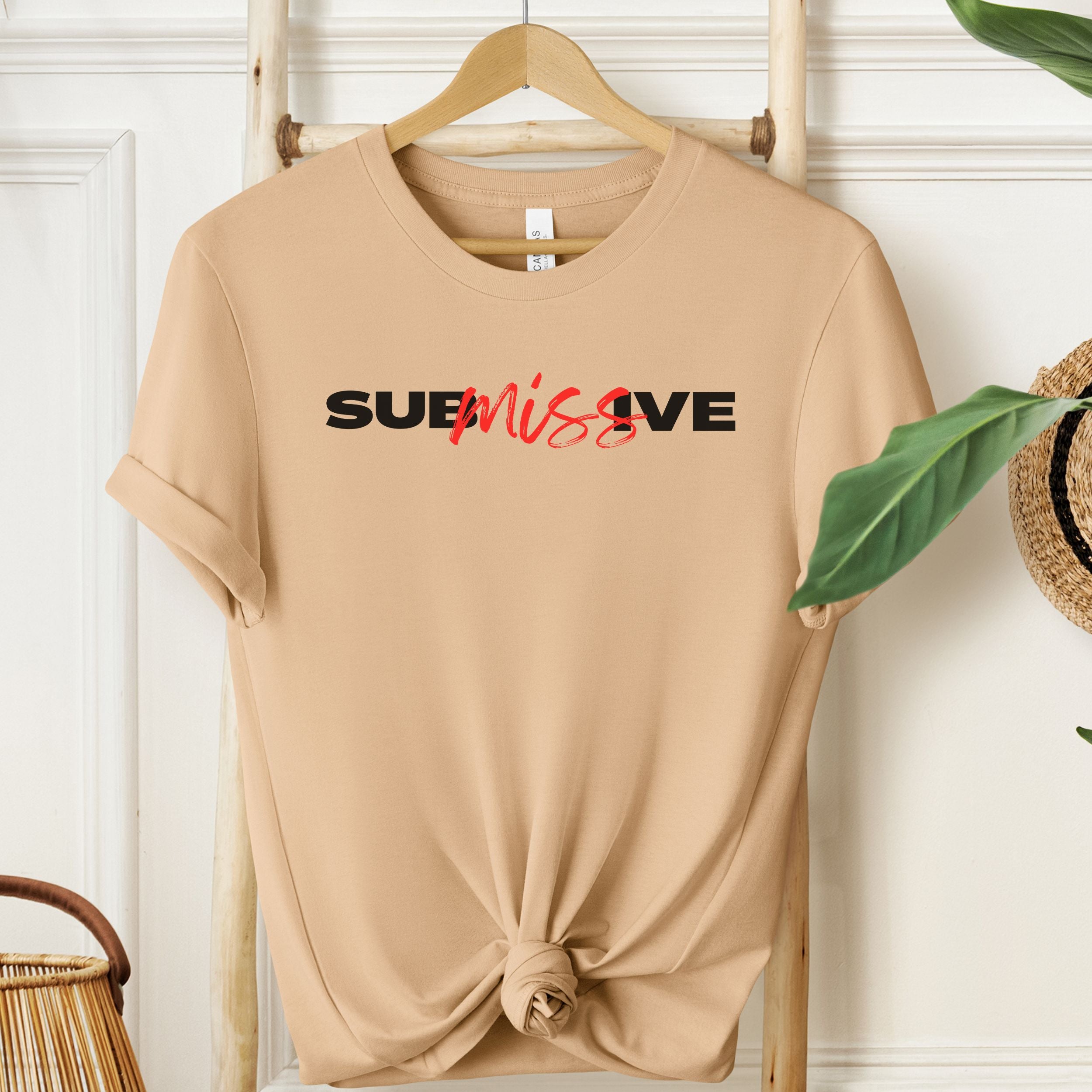 Sand Dune t-shirt with 'SUBmissive' text on a wooden hanger against a white wall.