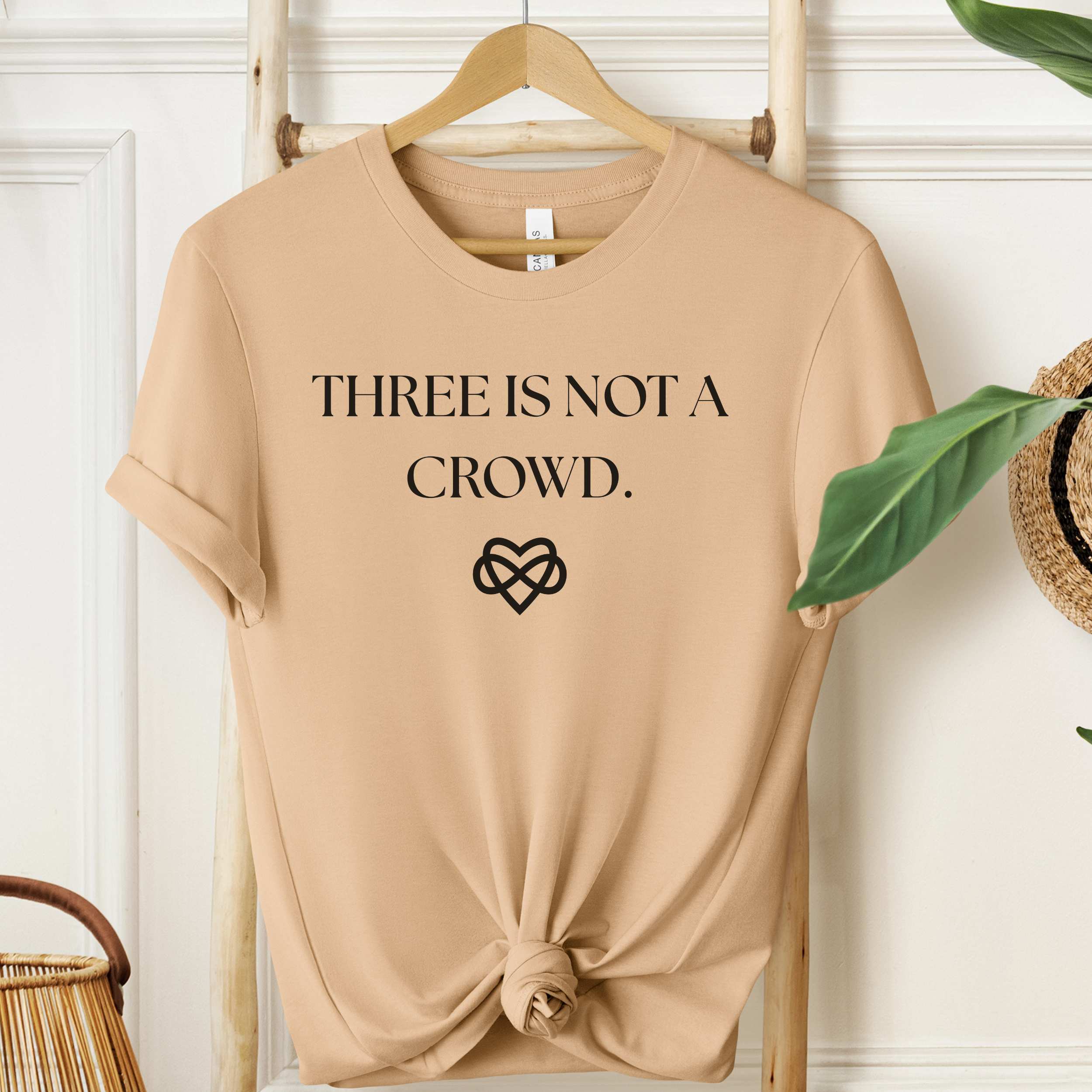 Beige t-shirt with 'THREE IS NOT A CROWD.' text and heart design on a wooden hanger.