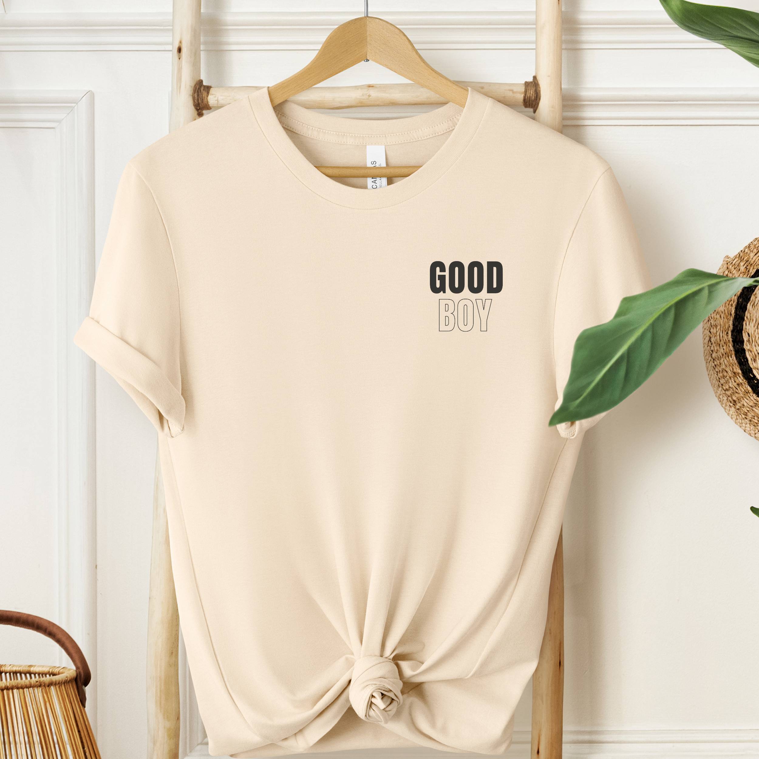 Beige t-shirt with 'GOOD BOY' text on a wooden hanger against a white wall.