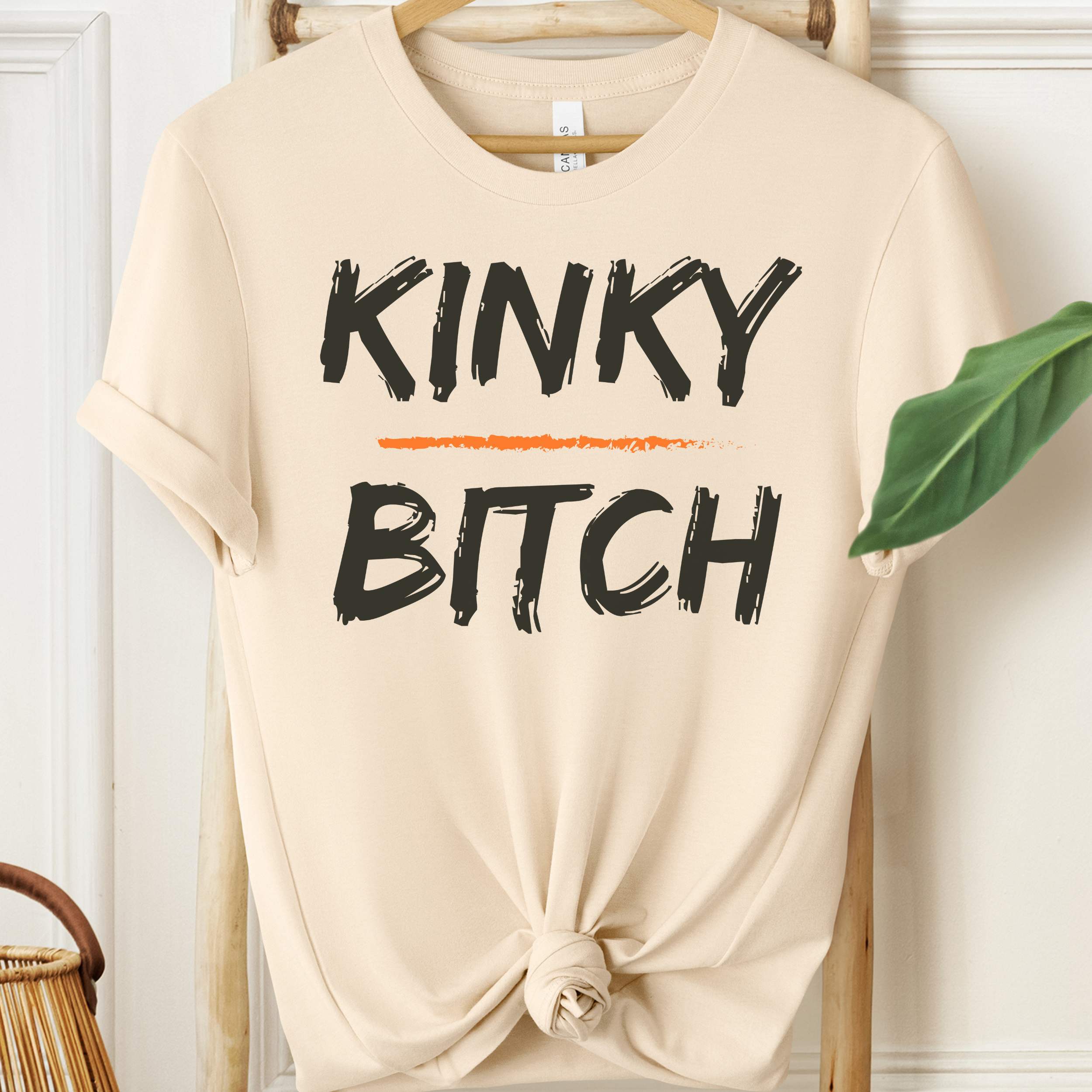 Beige t-shirt with 'KINKY BITCH' text on a hanger against a white wall.