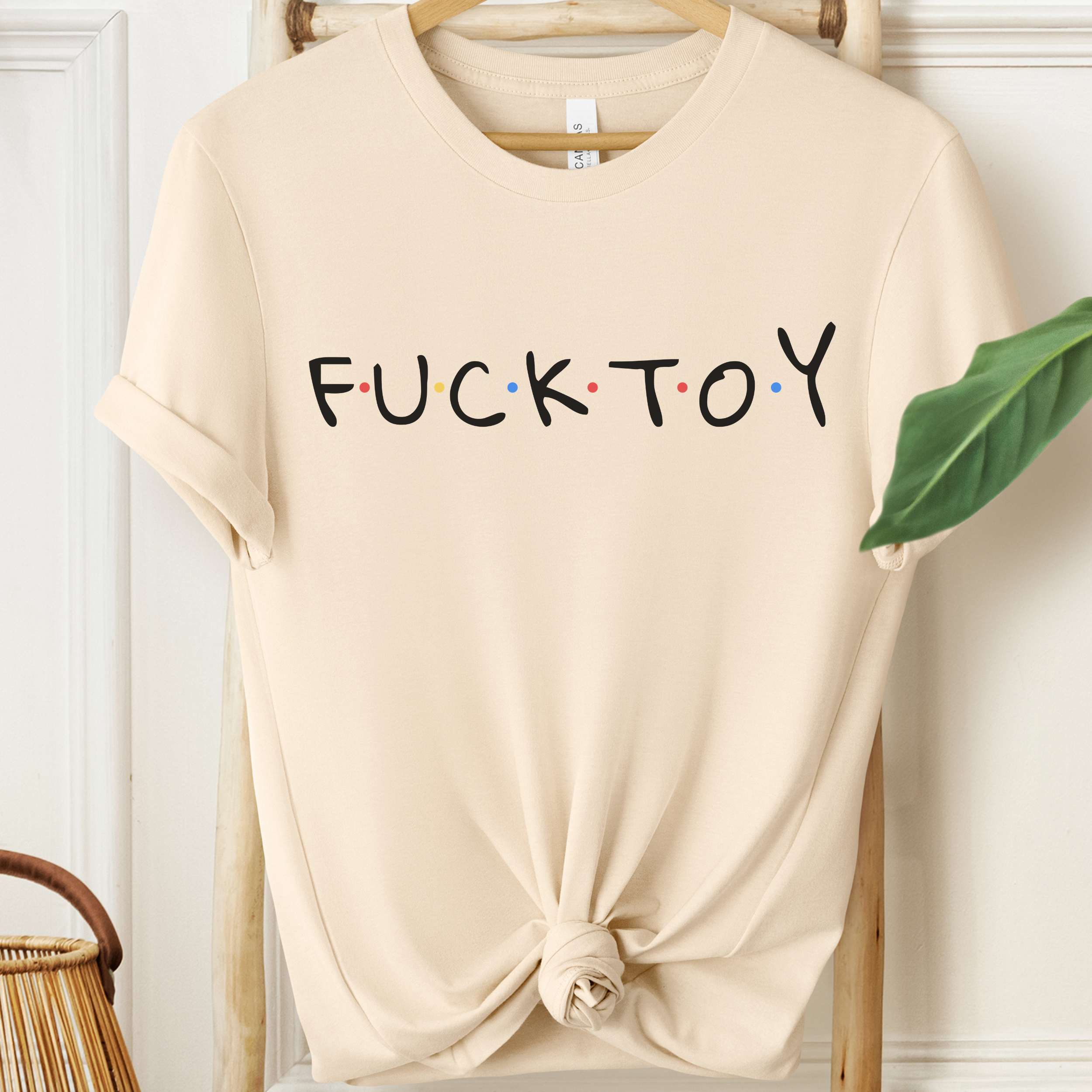 Beige t-shirt with 'FUCK TOY' text on a hanger against a white wall.