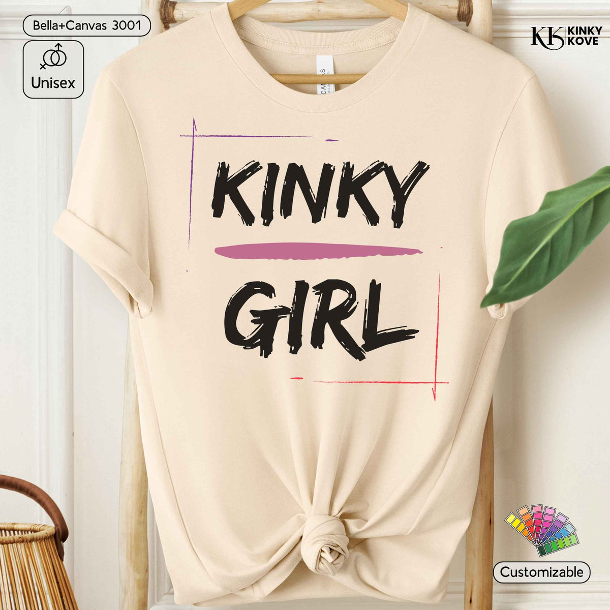 A beige short-sleeve t-shirt with a knotted hem, featuring the text 'KINKY GIRL' in black brush lettering with a pink underline and thin red and purple graphic lines. The shirt is displayed on a wooden hanger against a light background with decorative props.