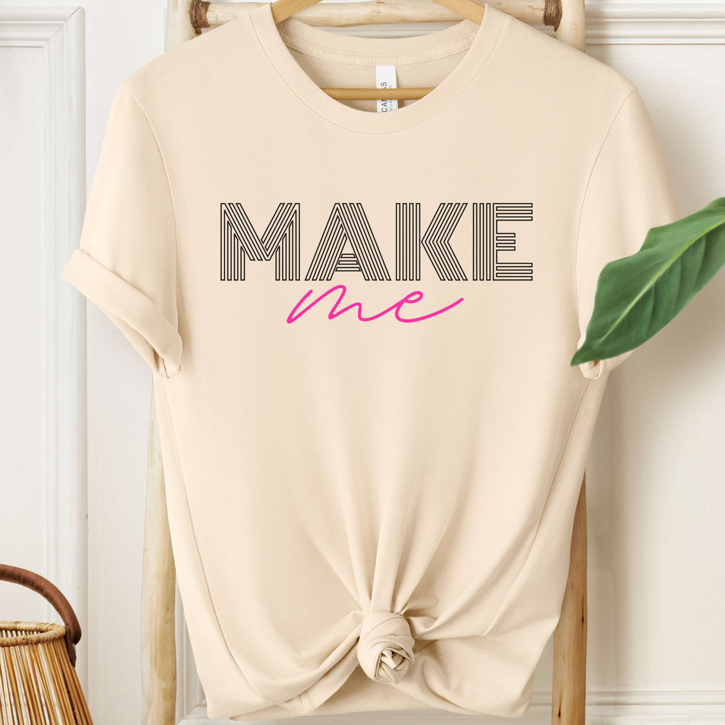 Beige t-shirt with 'MAKE me' text on a hanger against a white wall.