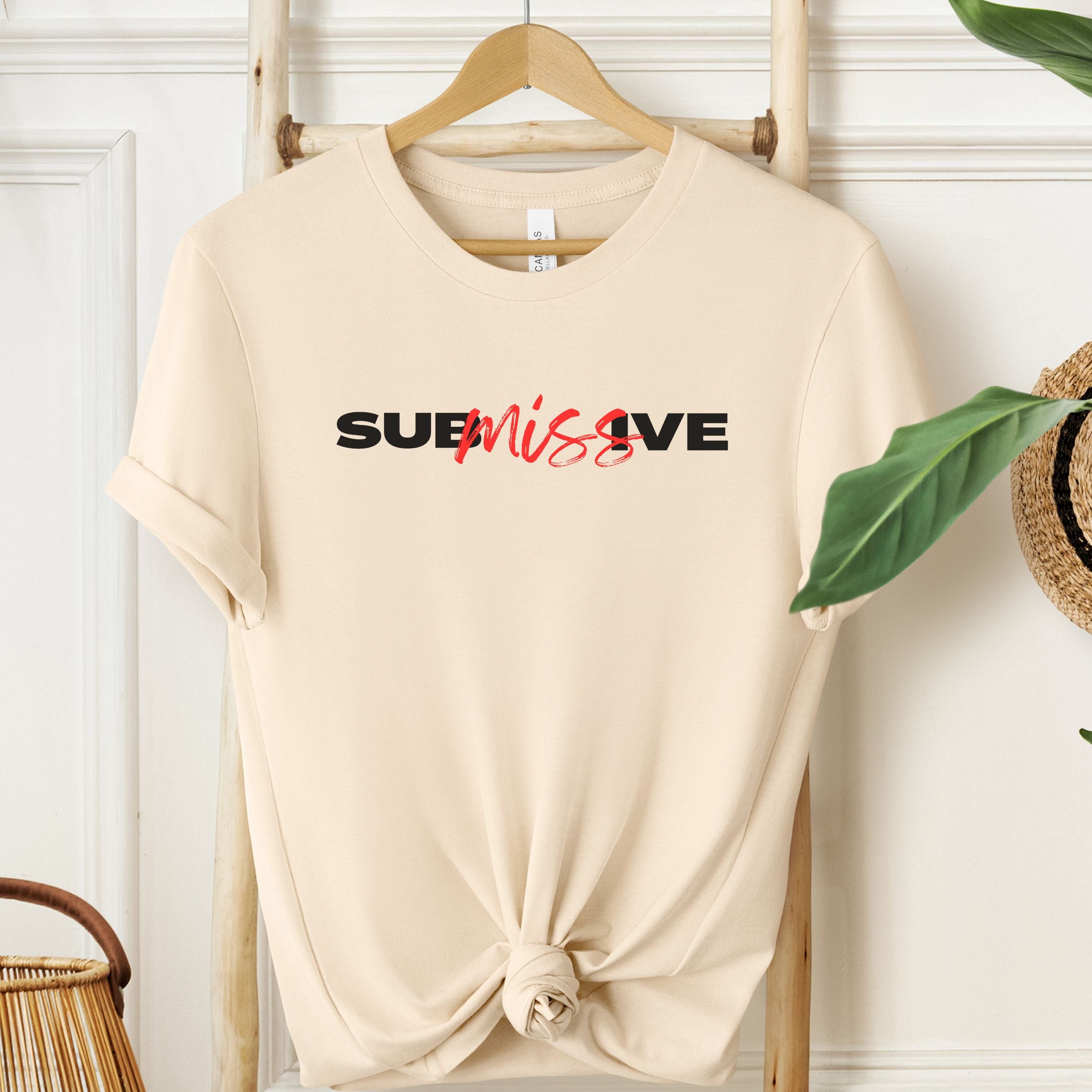 Beige t-shirt with 'SUBMISSIVE' text on a wooden hanger against a white wall.