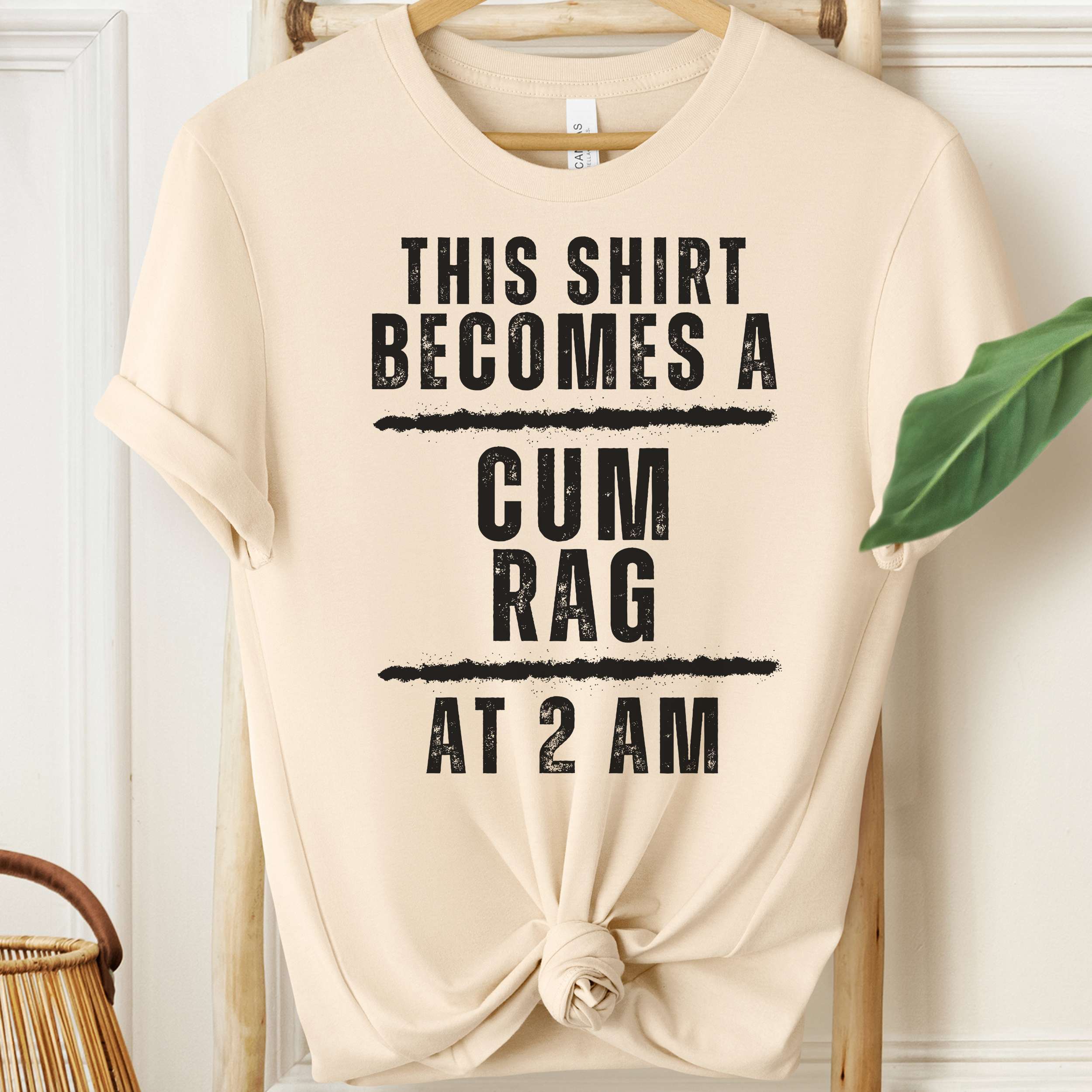 Beige t-shirt with black text on a hanger against a white wall.
