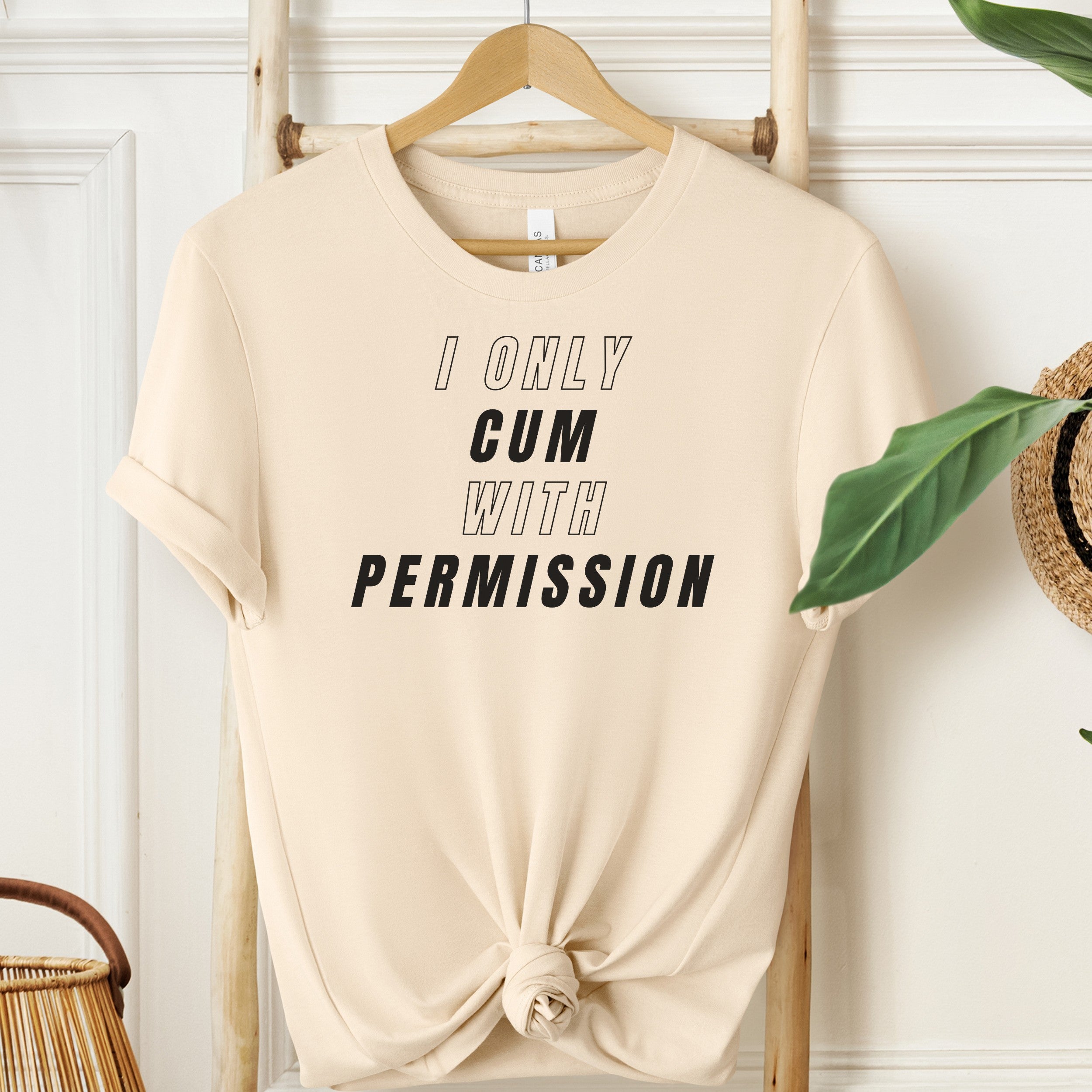 Beige t-shirt with text 'I ONLY GUM WITH PERMISSION' on a wooden hanger against a white wall.