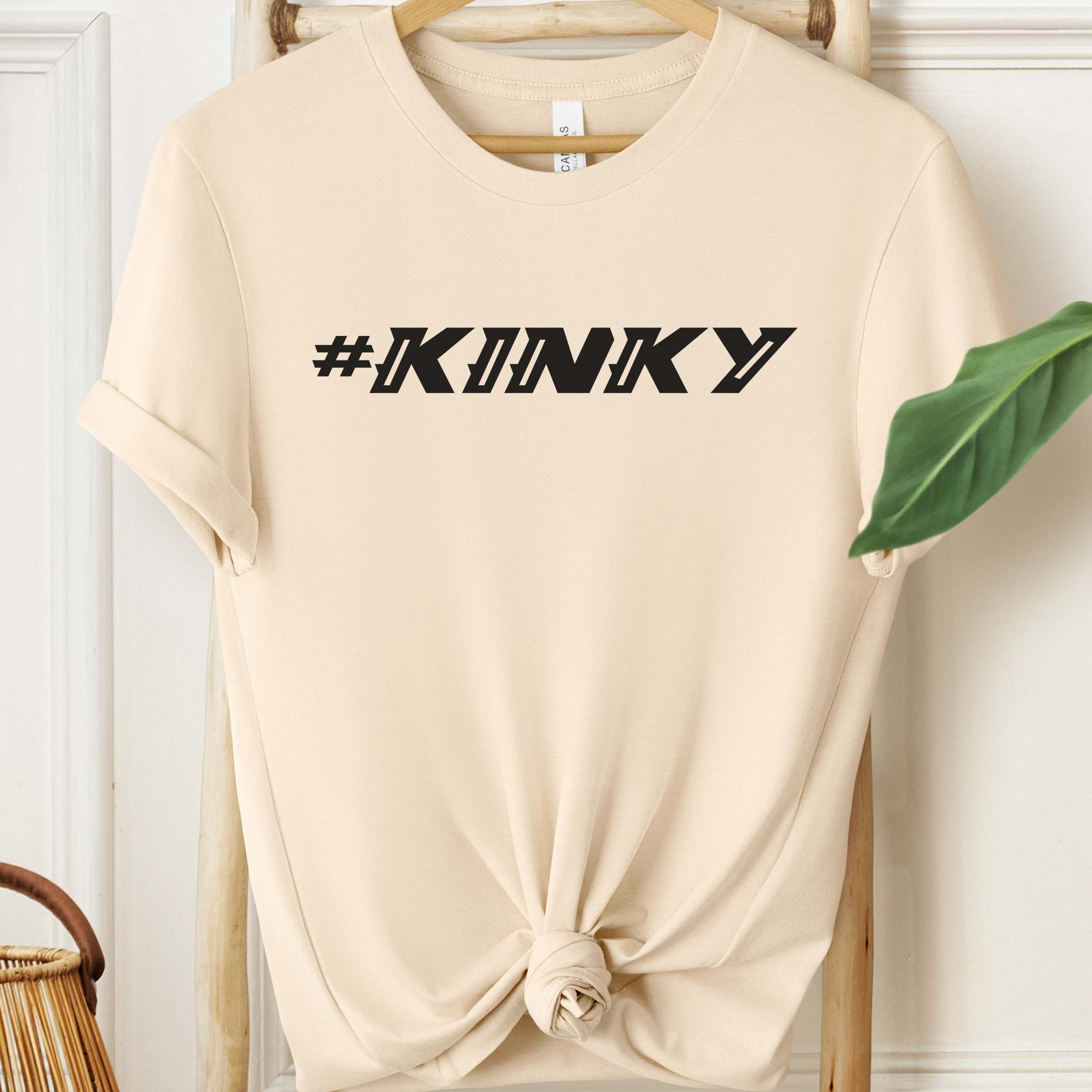 Beige t-shirt with '#KINKY' text on a hanger against a white wall.