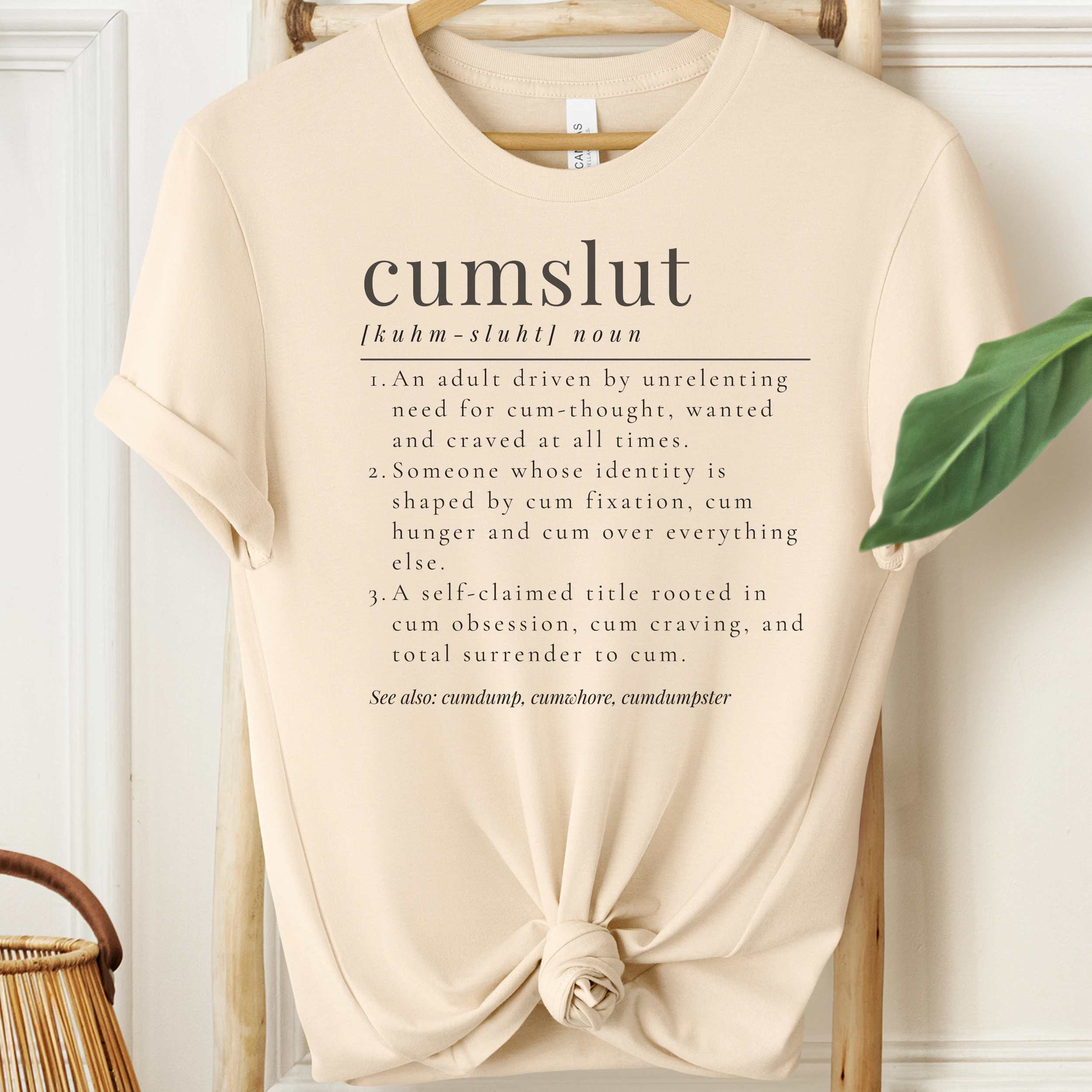 Beige t-shirt with text design on a hanger against a white wall.