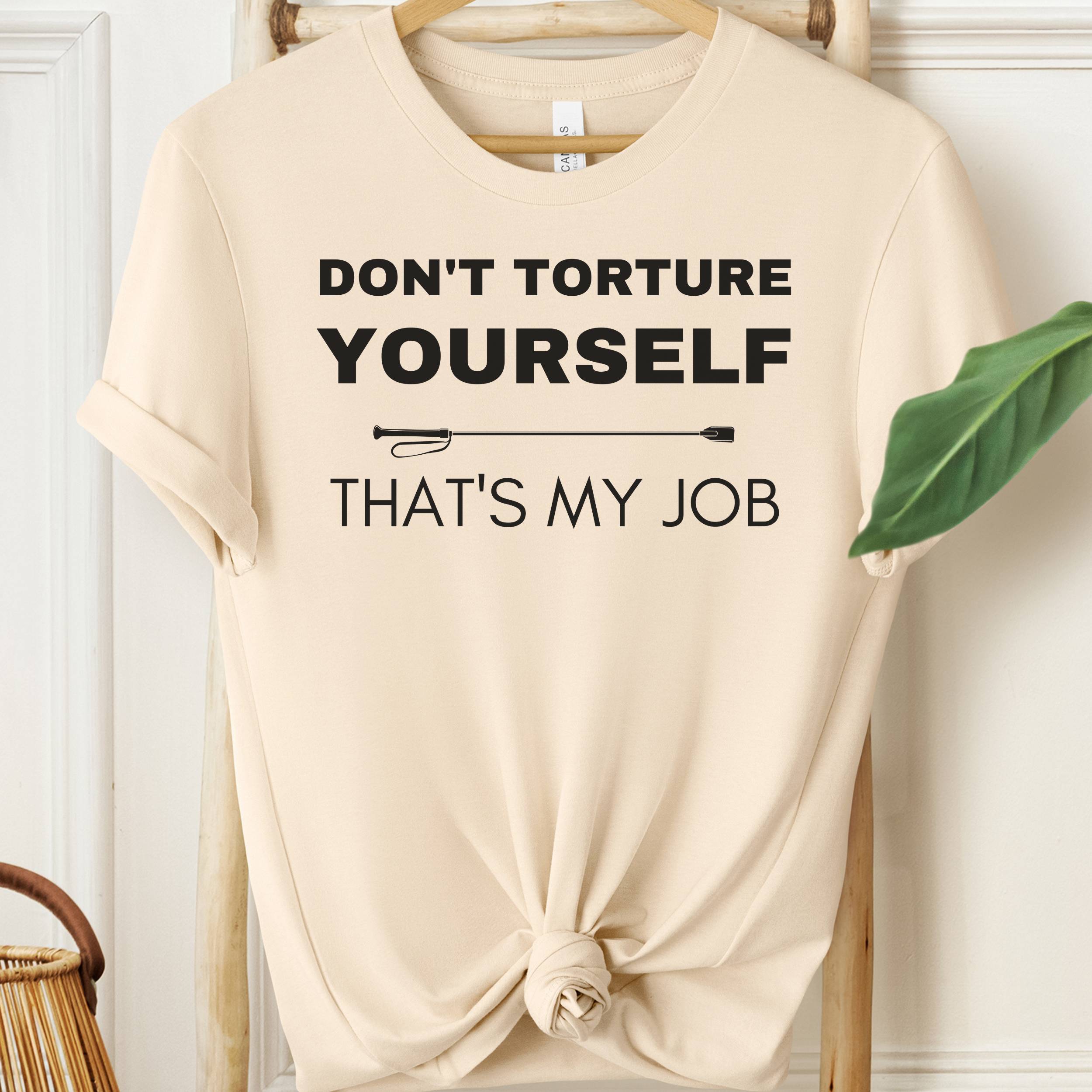 Beige t-shirt with text 'Don't torture yourself, that's my job' on a hanger.