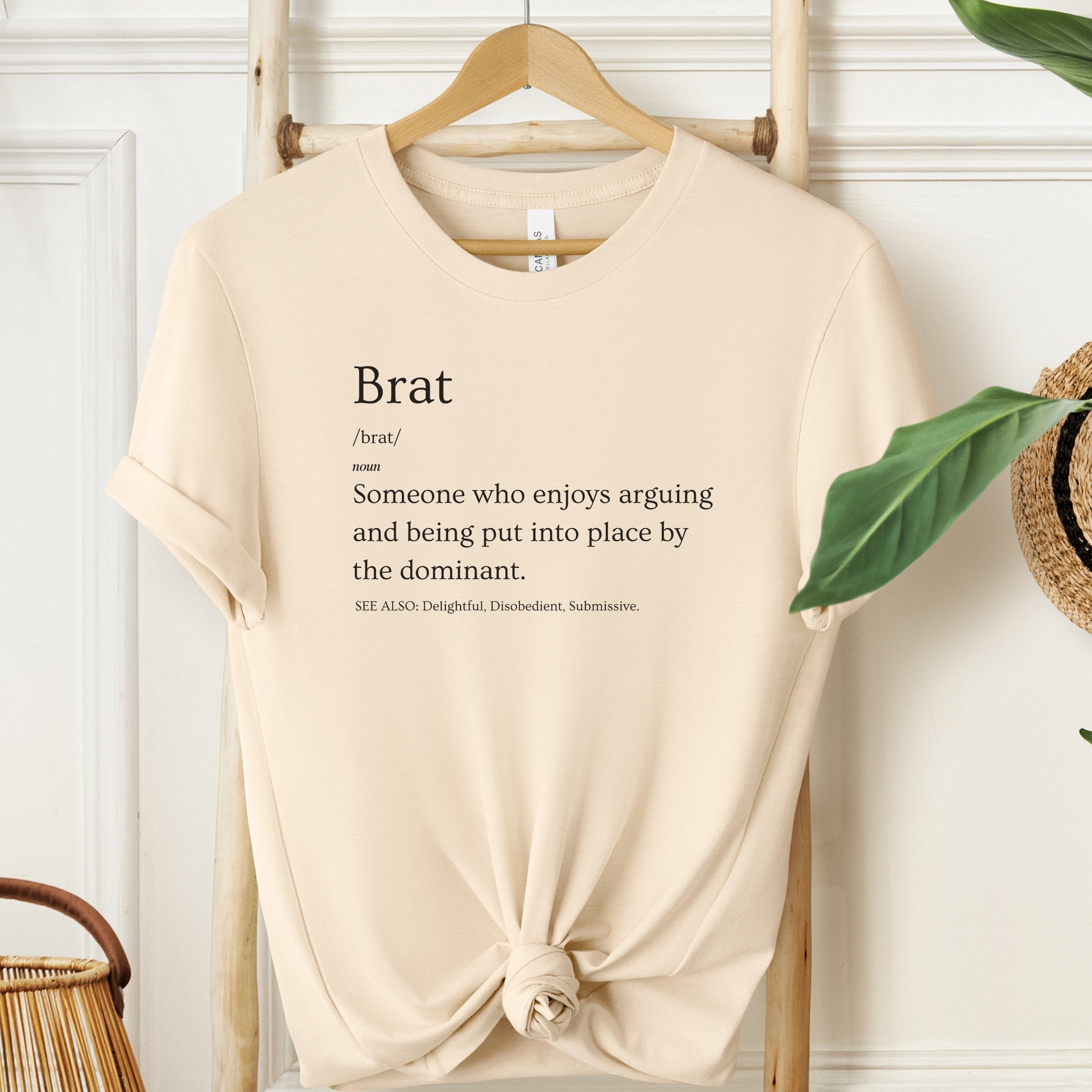 Beige t-shirt with printed definition of 'brat' on a wooden hanger against a white wall.