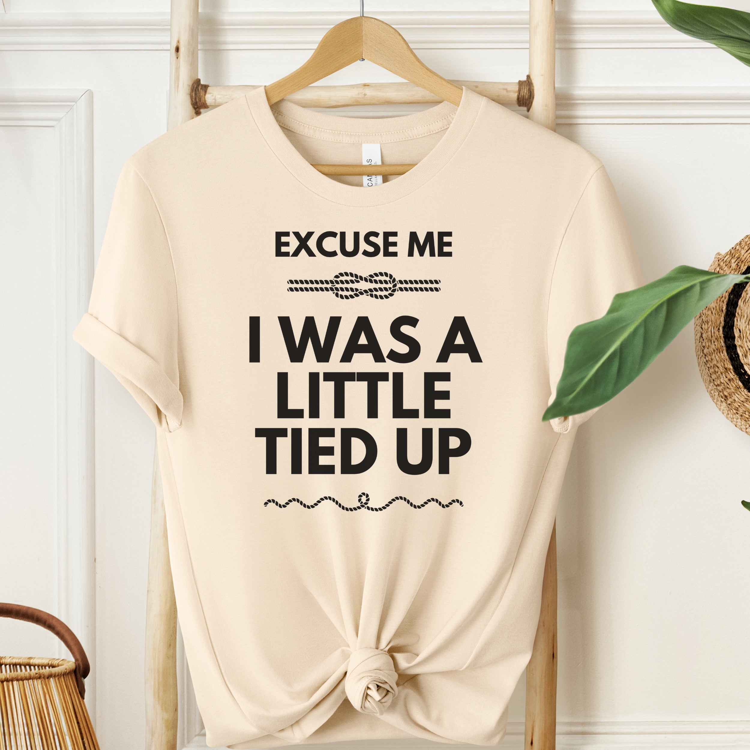 Beige t-shirt with text 'Excuse me I was a little tied up' on a wooden hanger.