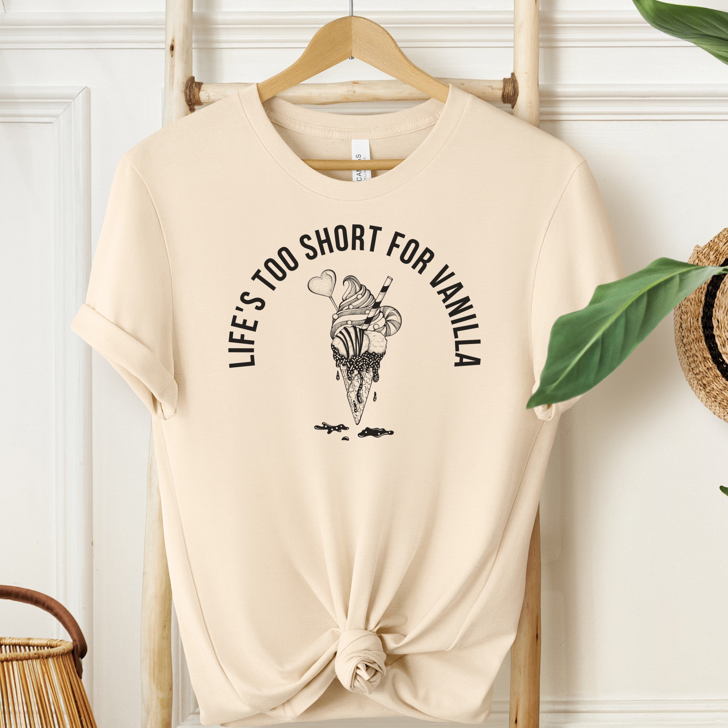 Beige t-shirt with ice cream graphic and text on a wooden hanger against a white wall.