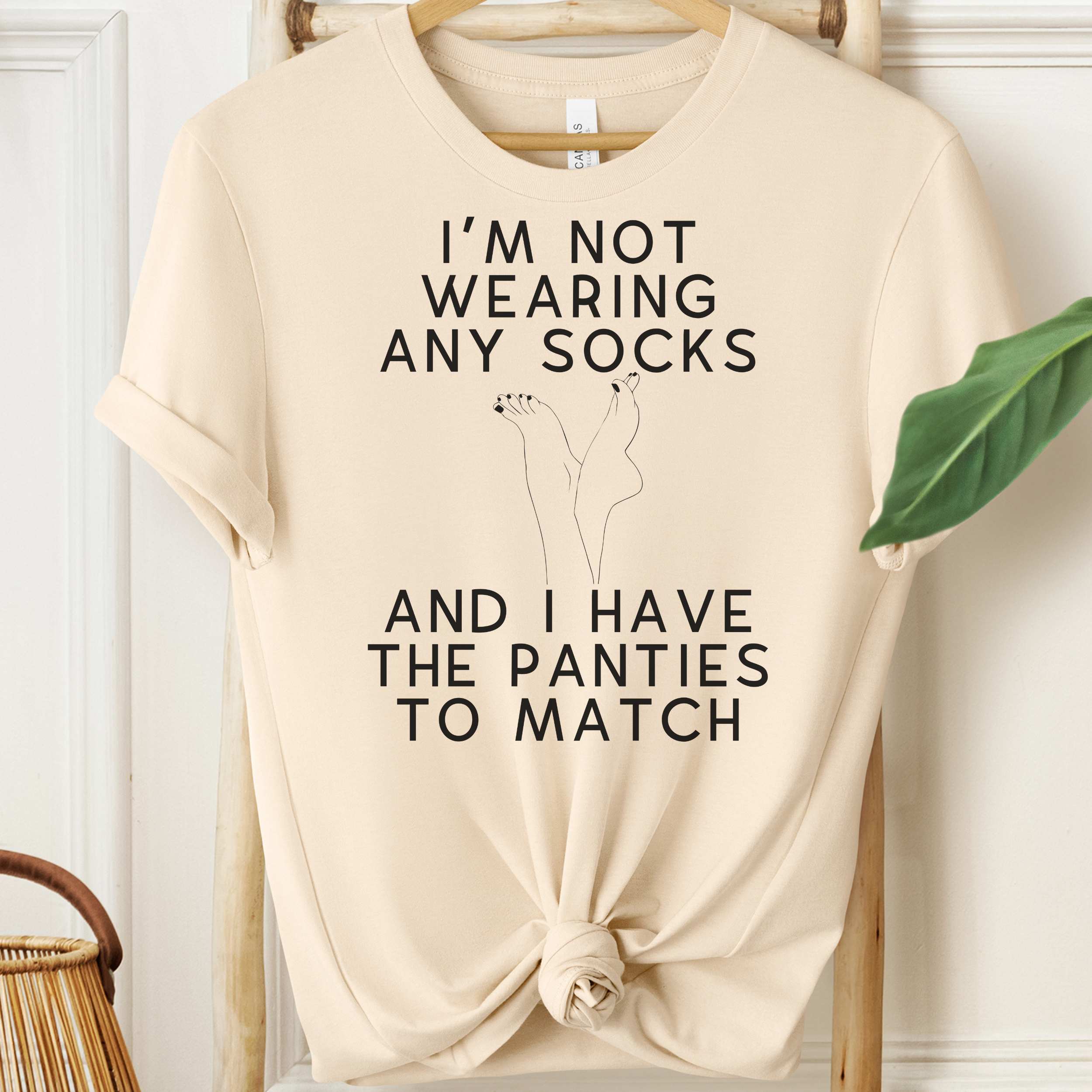 Beige t-shirt with humorous text design on a hanger against a white wall.