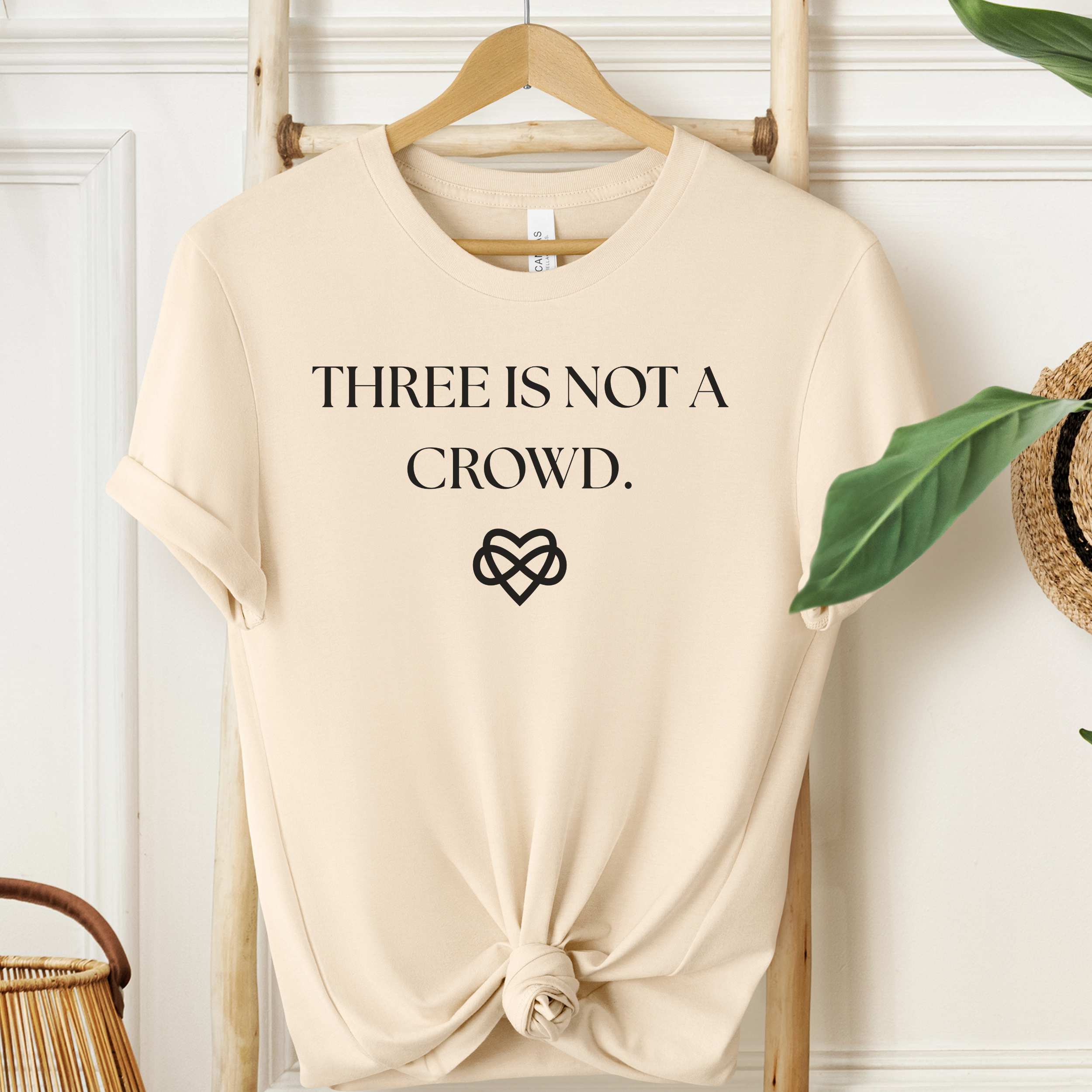 Beige t-shirt with 'THREE IS NOT A CROWD.' text and heart symbol on a wooden hanger.