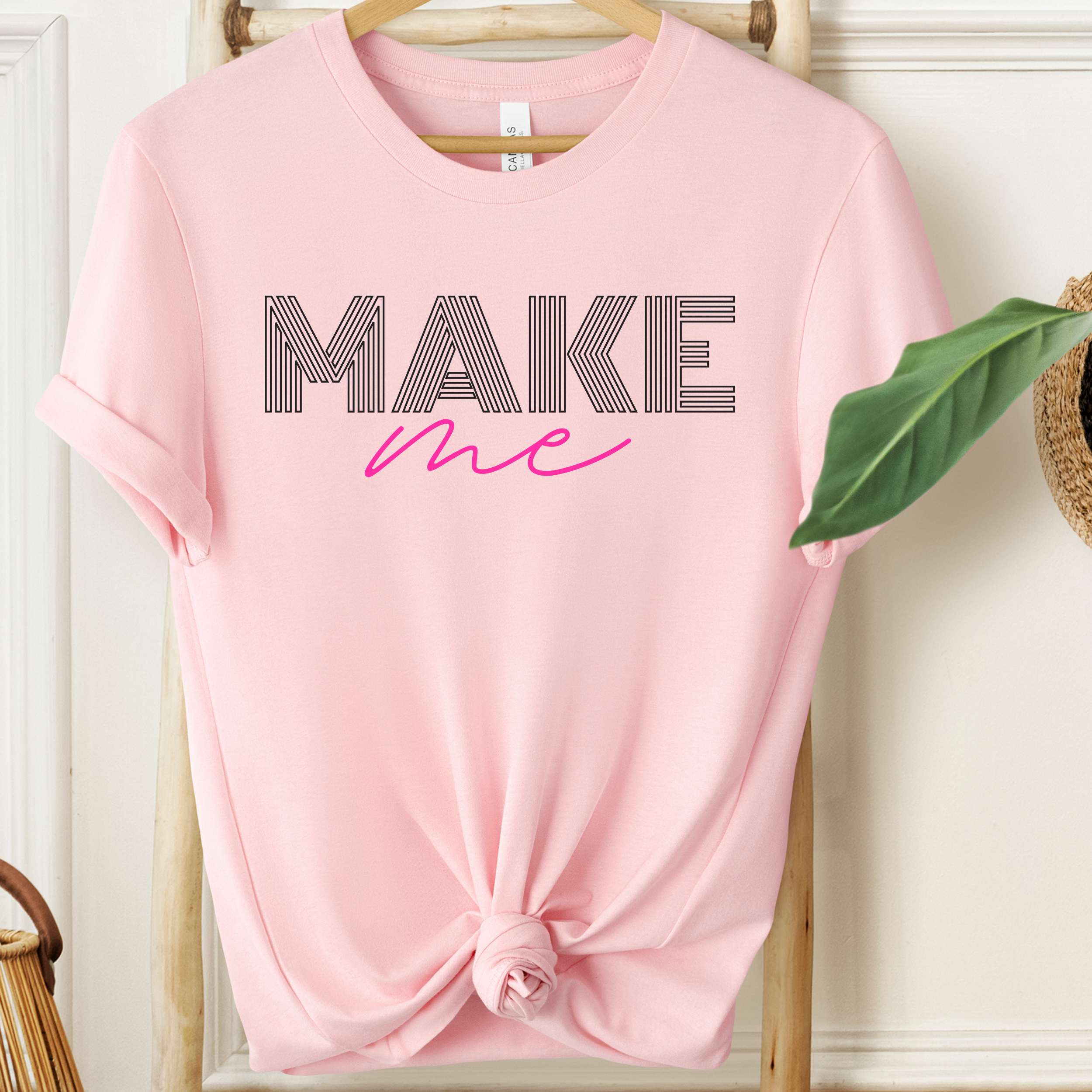 Pink t-shirt with 'MAKE me' text on a wooden hanger.