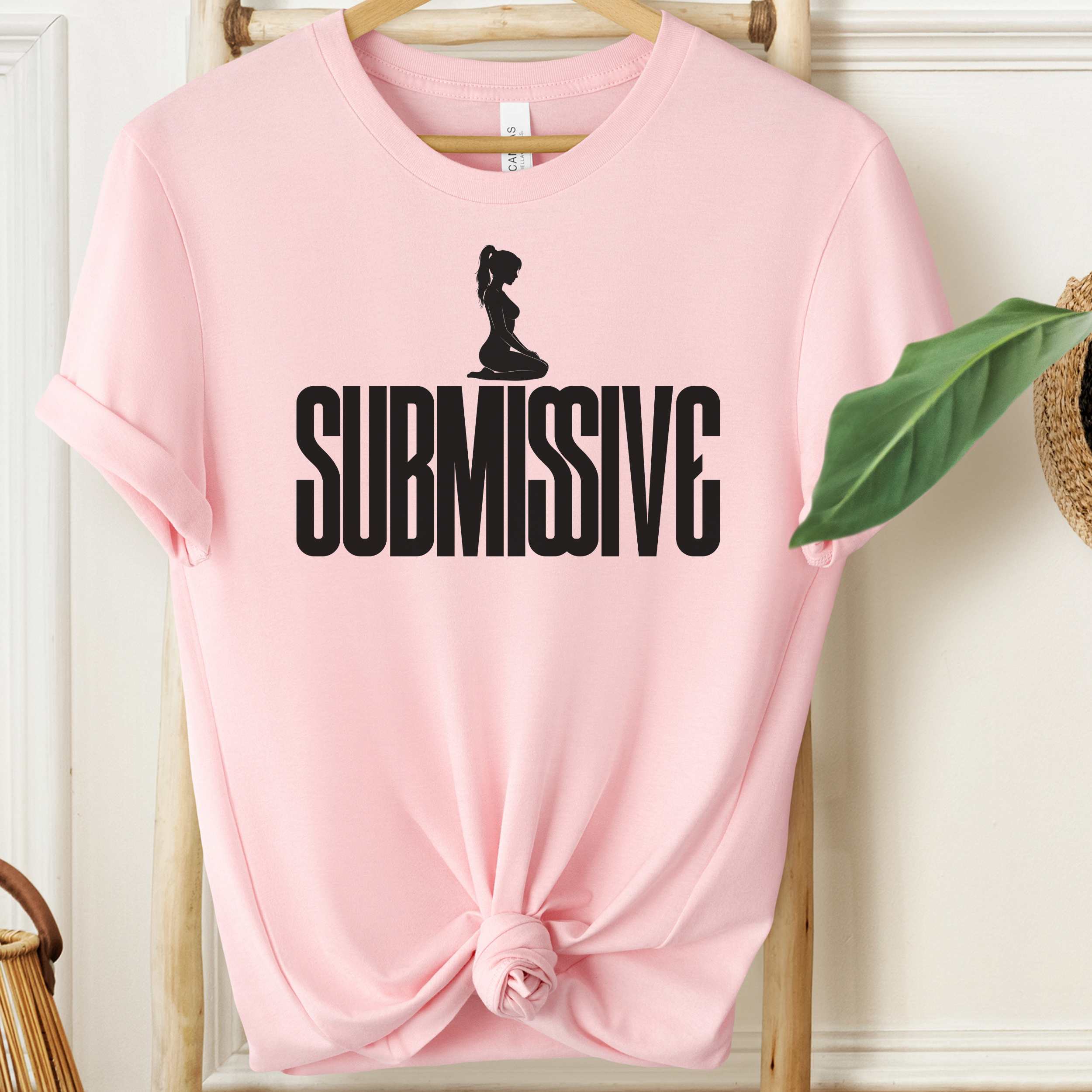 Pink t-shirt with 'SUBMISSIVE' text and silhouette graphic on a wooden hanger.