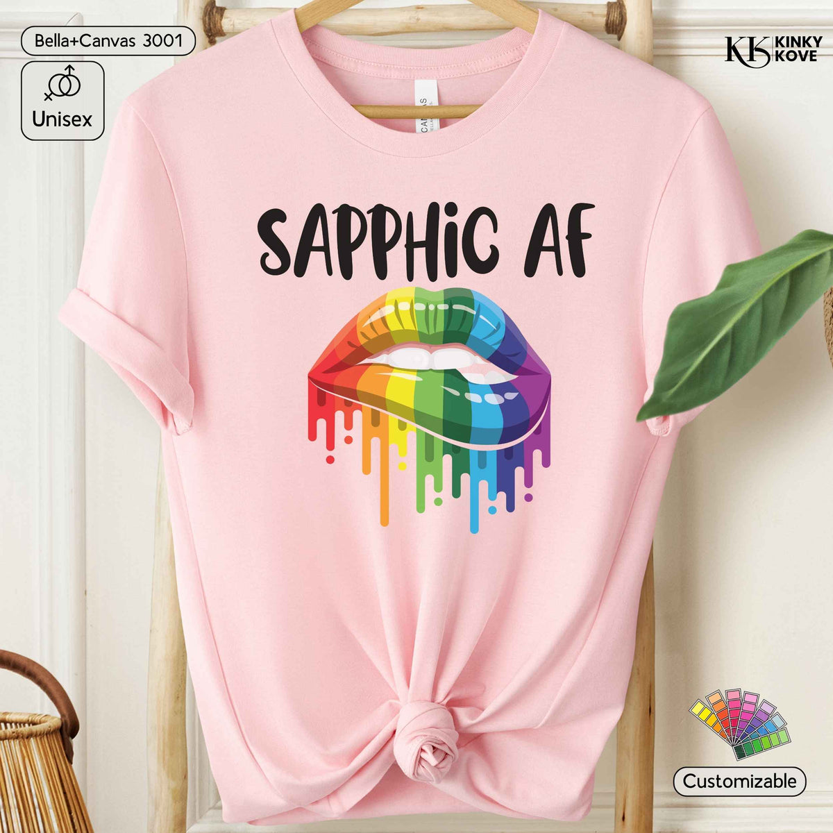 A light pink short-sleeve t-shirt with a graphic of dripping rainbow-colored lips and the text 'SAPPHIC AF' in black. The t-shirt is displayed on a wooden hanger against a light-colored wall with decorative props.
