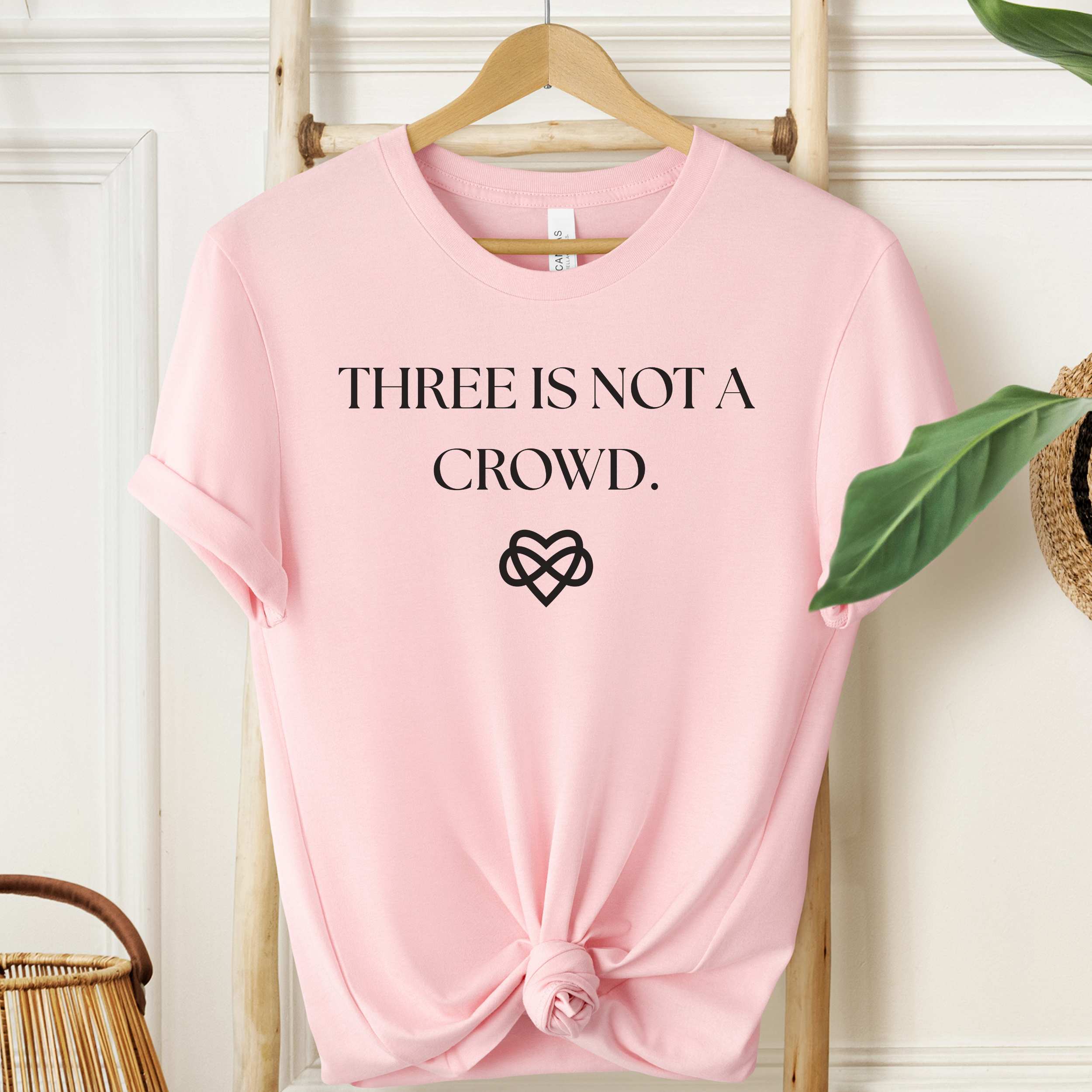 Pink t-shirt with 'THREE IS NOT A CROWD.' text and heart symbol on a hanger.