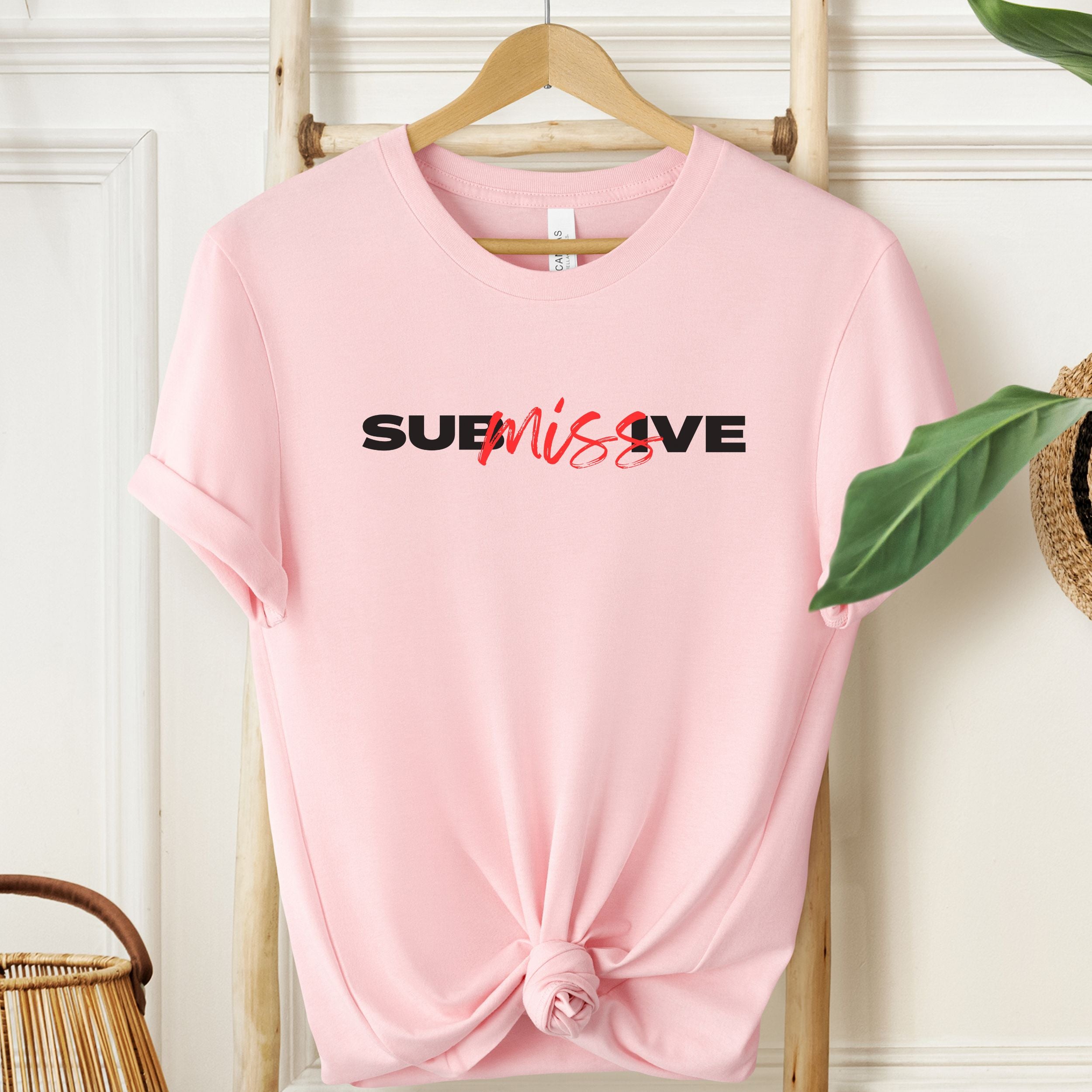 Pink t-shirt with 'SUBMISSIVE' text on a hanger against a white wall.