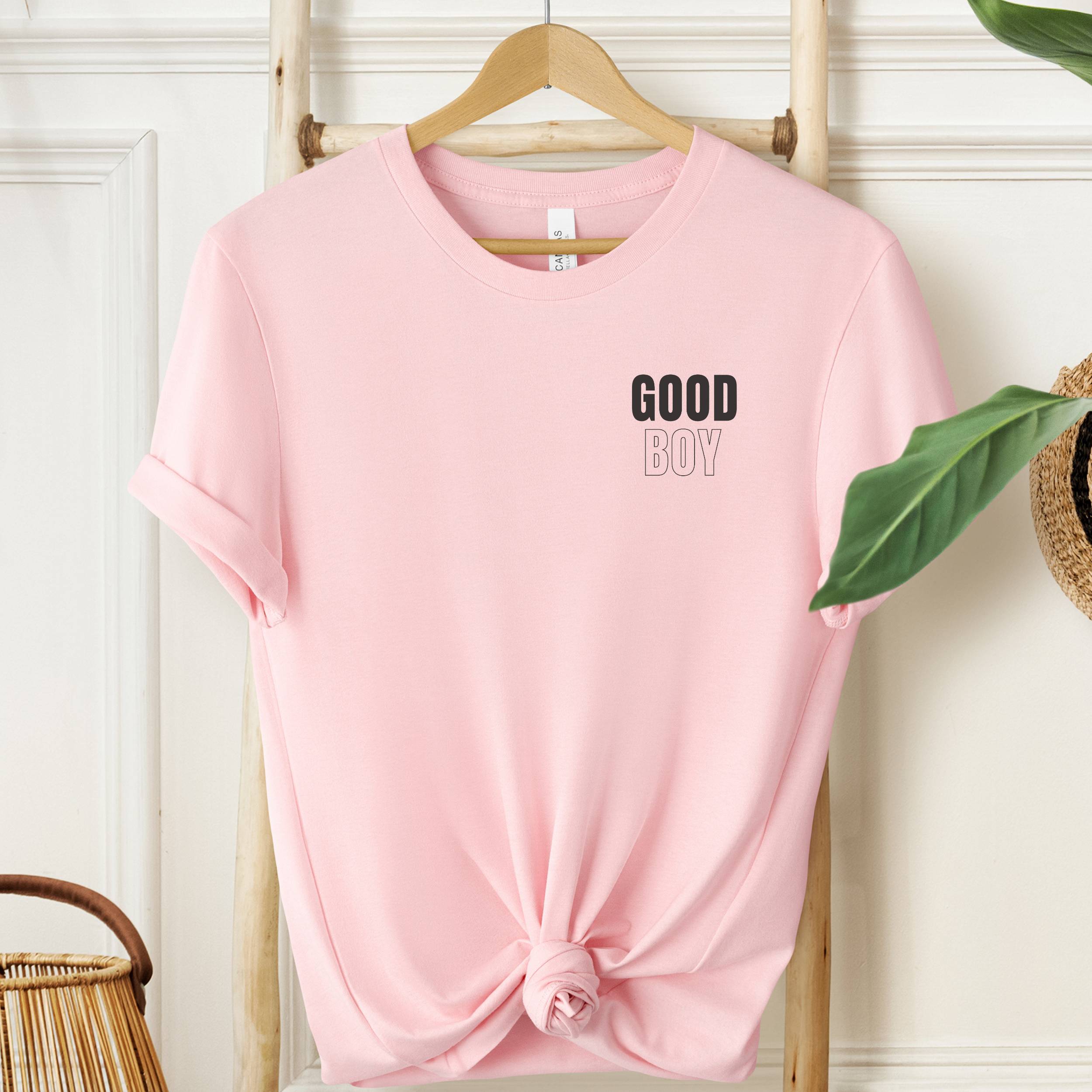 Pink t-shirt with 'GOOD BOY' text on a hanger against a white wall.