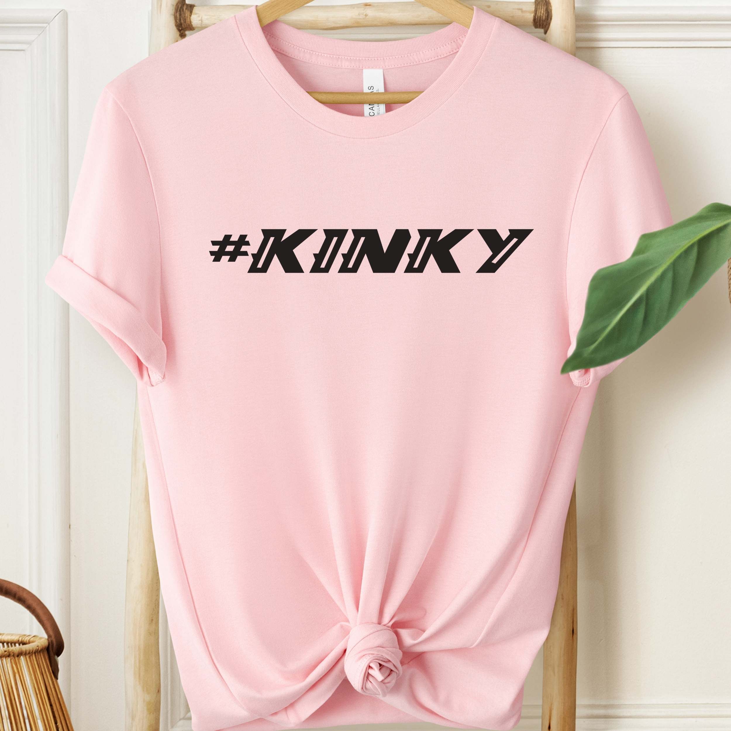 Pink t-shirt with '#KINKY' text on a hanger against a white wall.