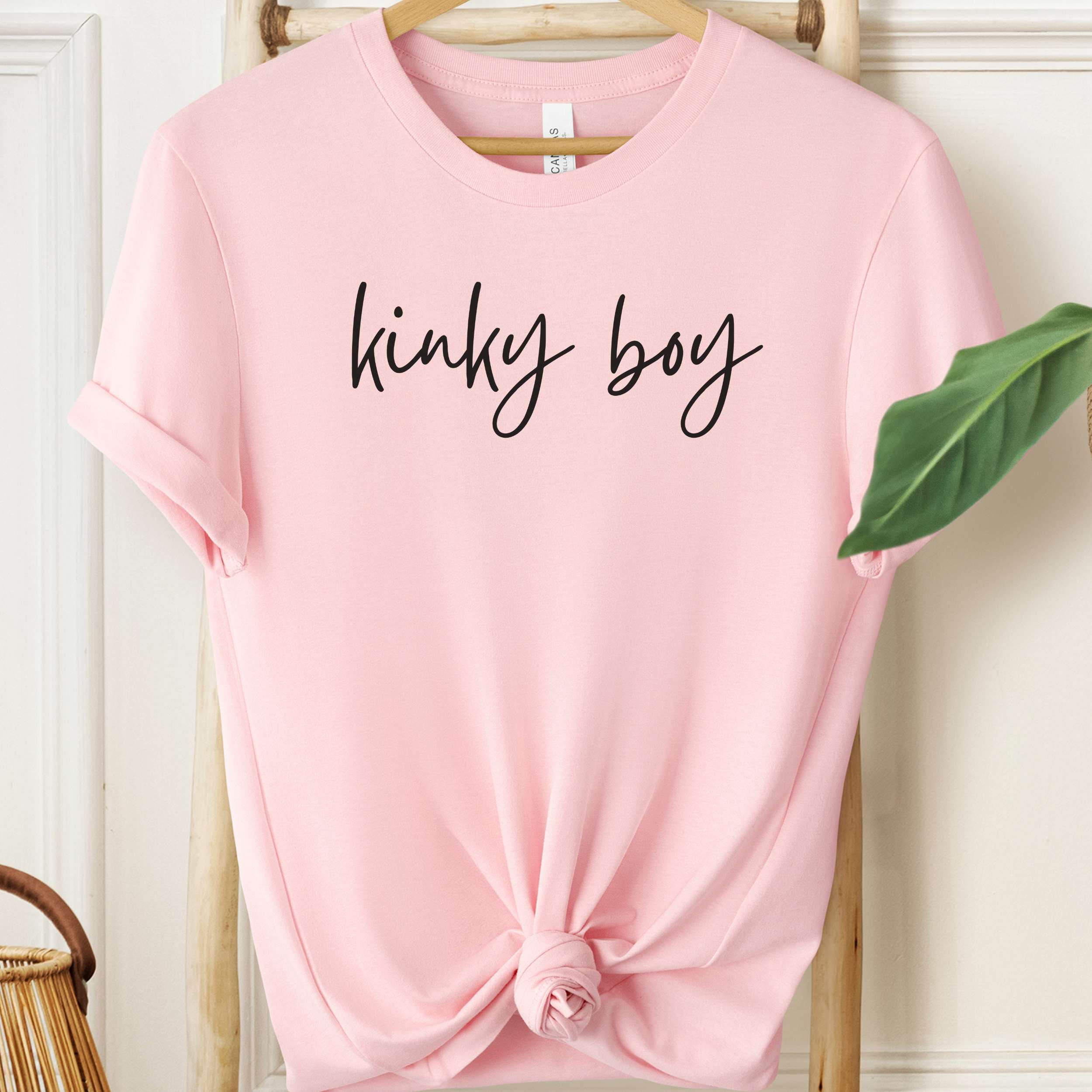 Pink t-shirt with 'kinky boy' text on a hanger against a white wall.