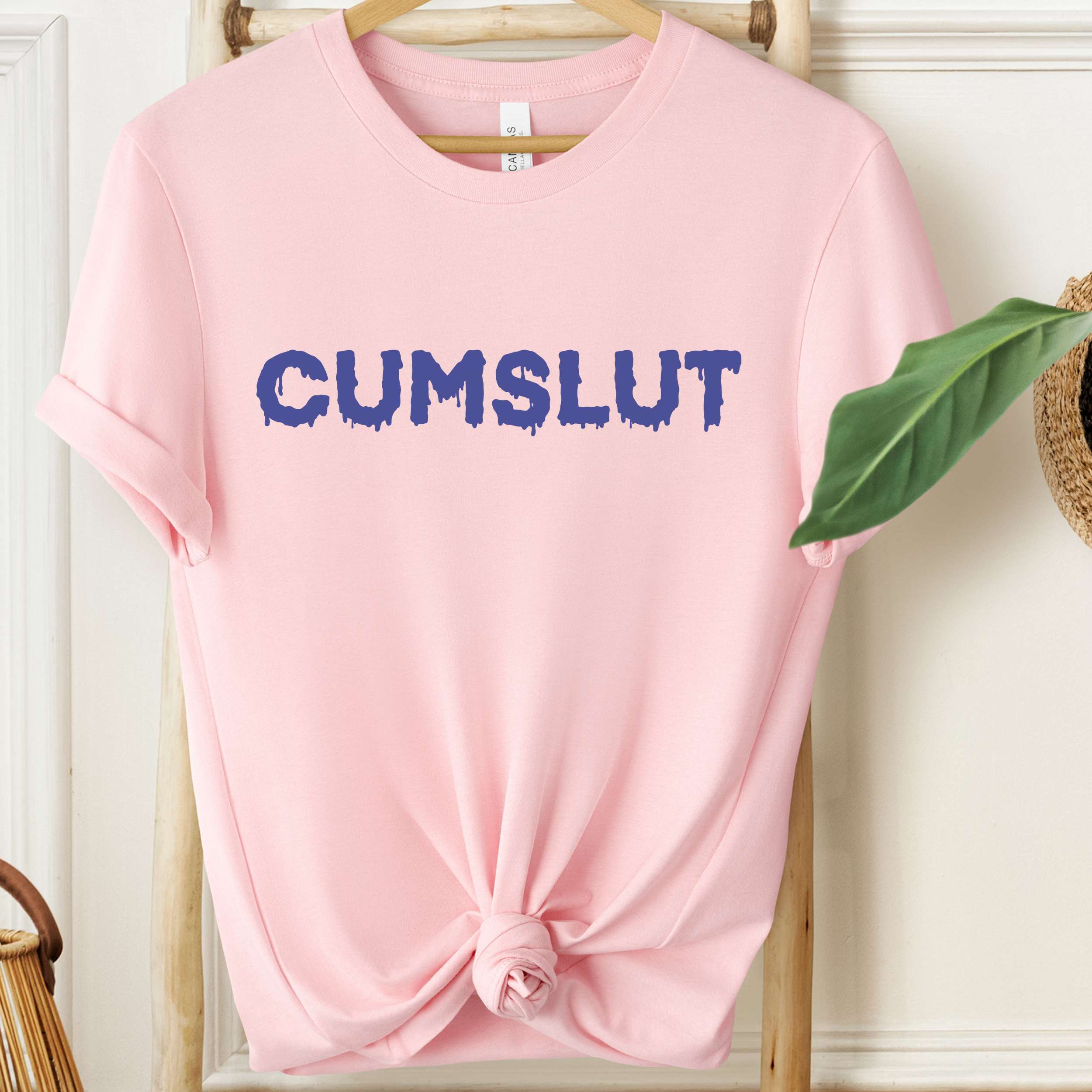 Pink t-shirt with 'CUMSLUT' printed in blue on a wooden hanger.