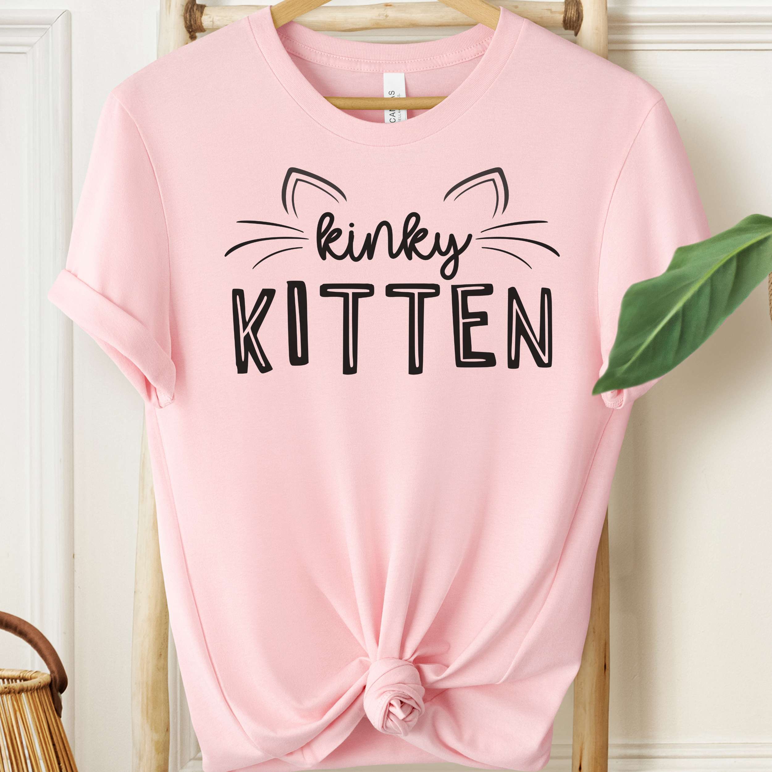 Pink t-shirt with 'kinky KITTEN' text on a hanger against a white wall.