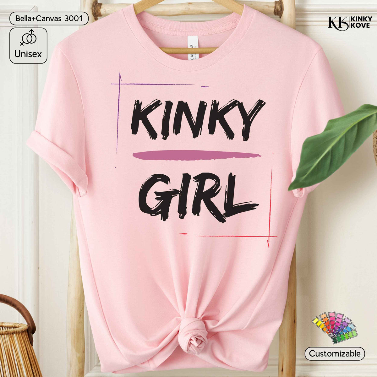 A pink short-sleeve t-shirt with a knot at the bottom hem, featuring the words 'KINKY GIRL' printed in black brush-style lettering with a purple horizontal stripe. The shirt is displayed on a wooden hanger against a light wall, with decorative props including a green leaf, a woven basket, and informational badges.