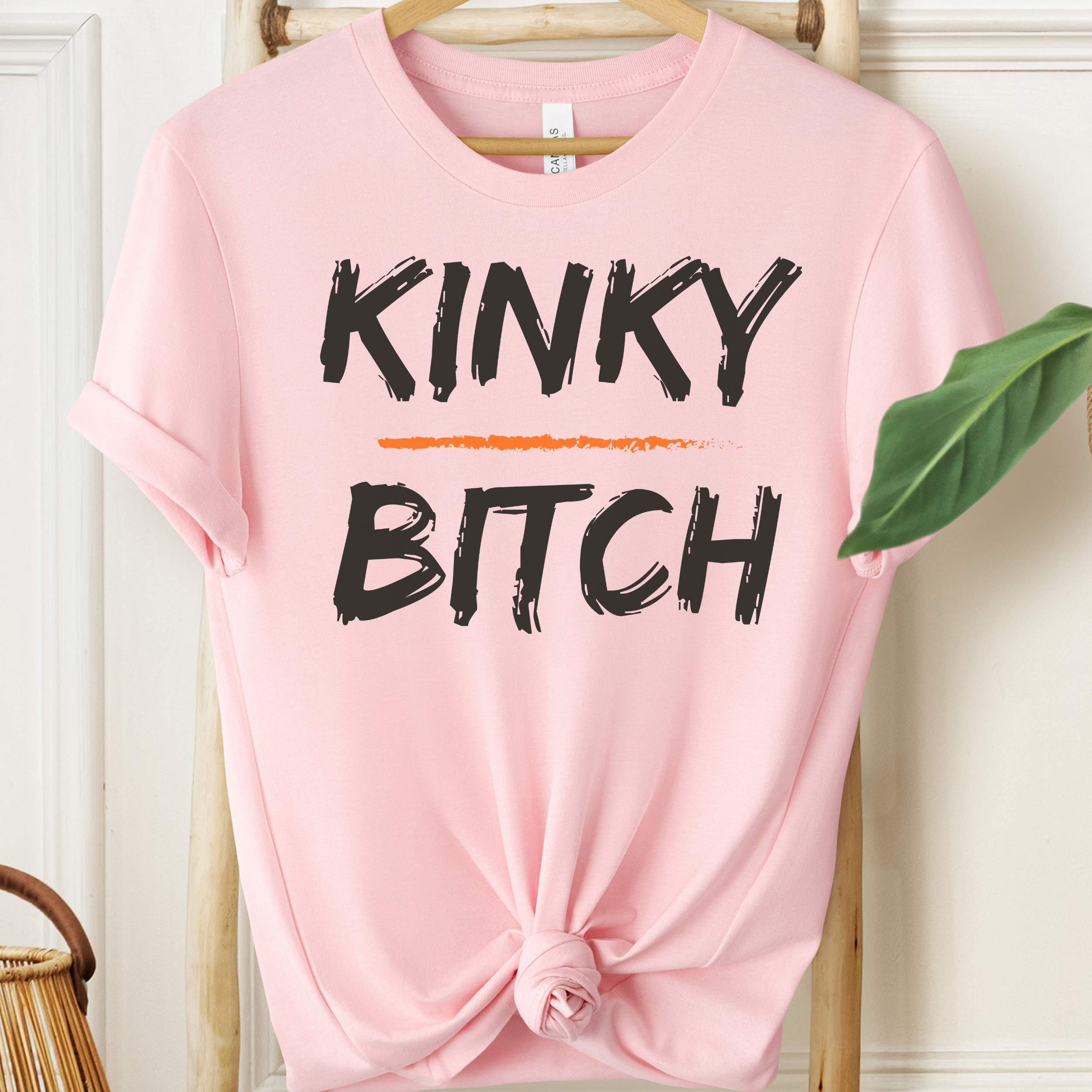 Pink t-shirt with 'KINKY BITCH' text on a hanger against a white wall.