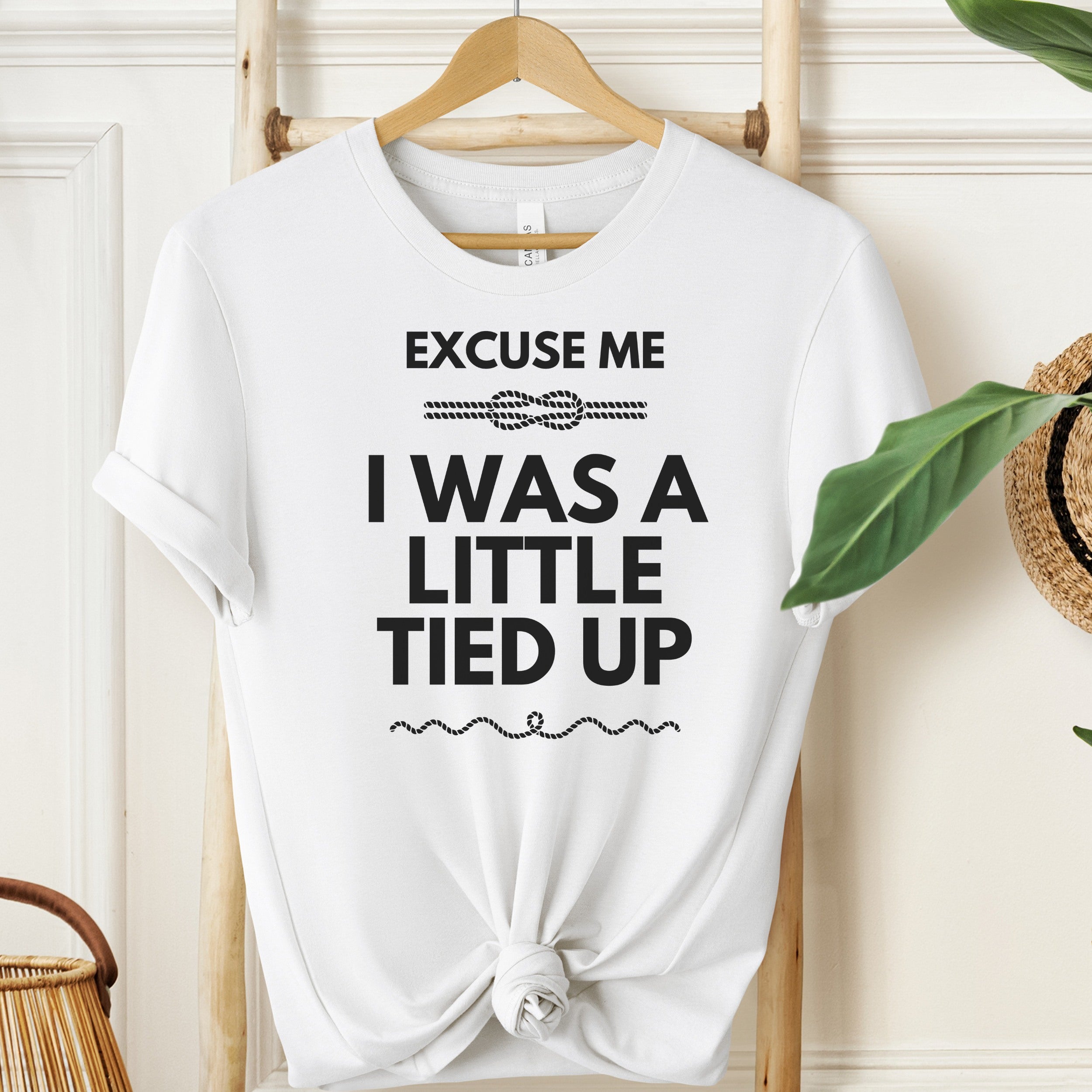 White t-shirt with text 'EXCUSE ME I WAS A LITTLE TIED UP' on a wooden hanger.