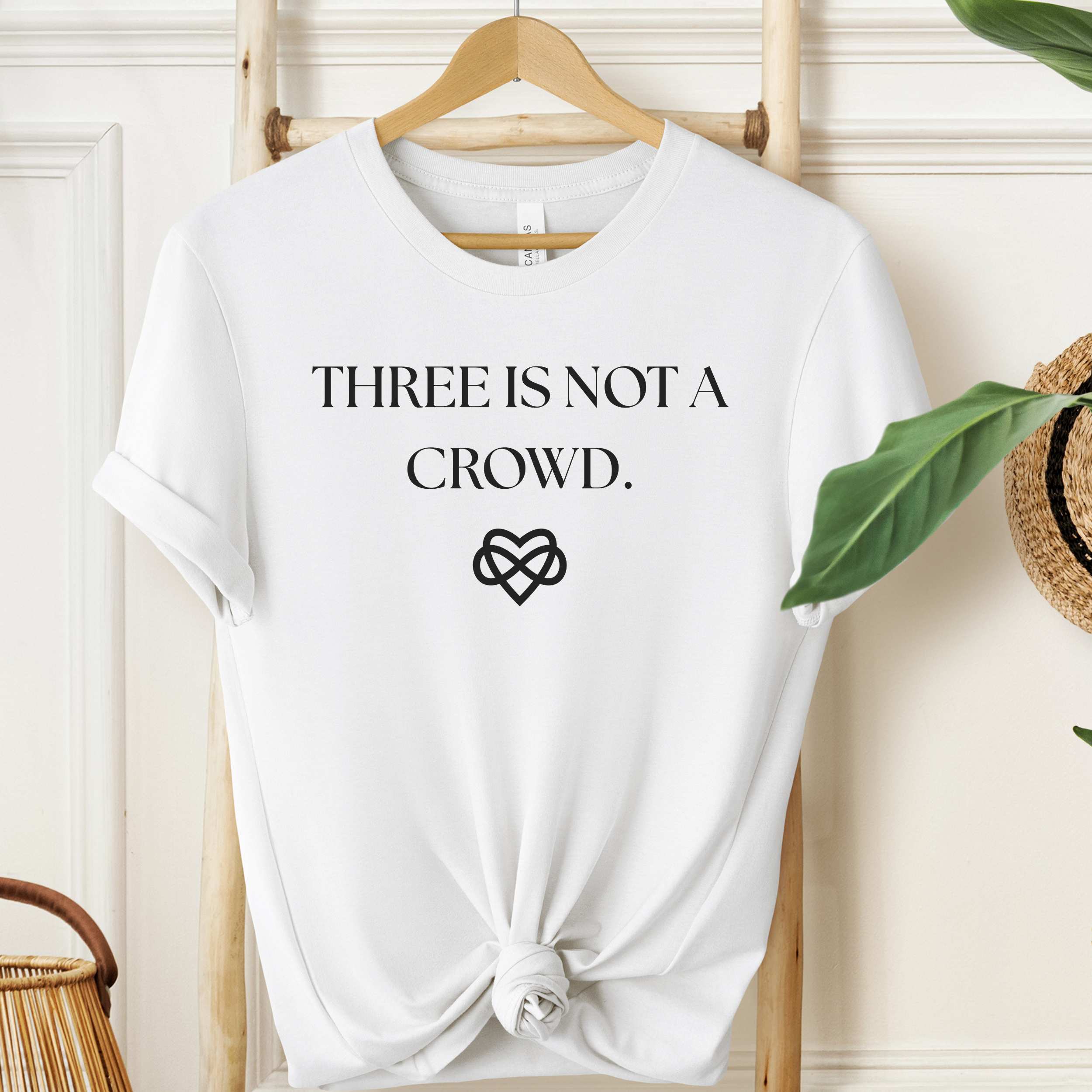 White t-shirt with 'THREE IS NOT A CROWD.' text and heart symbol on a wooden hanger.