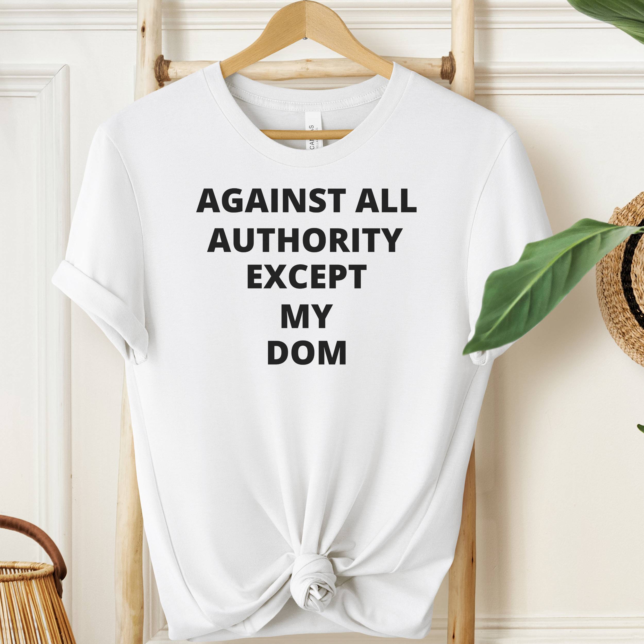 White t-shirt with text 'AGAINST ALL AUTHORITY EXCEPT MY DOM' on a hanger.