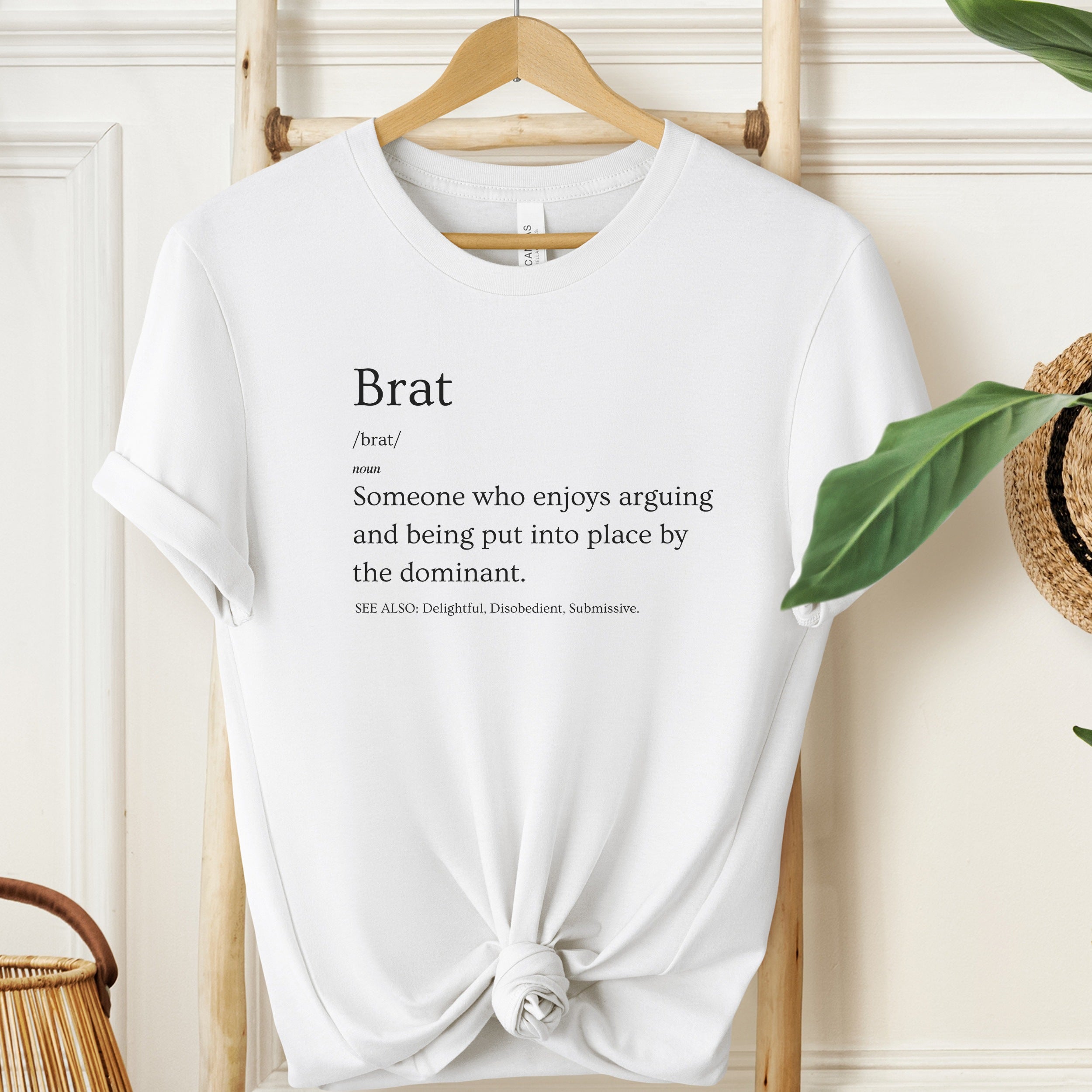 White t-shirt with 'brat' definition text on a hanger against a neutral background