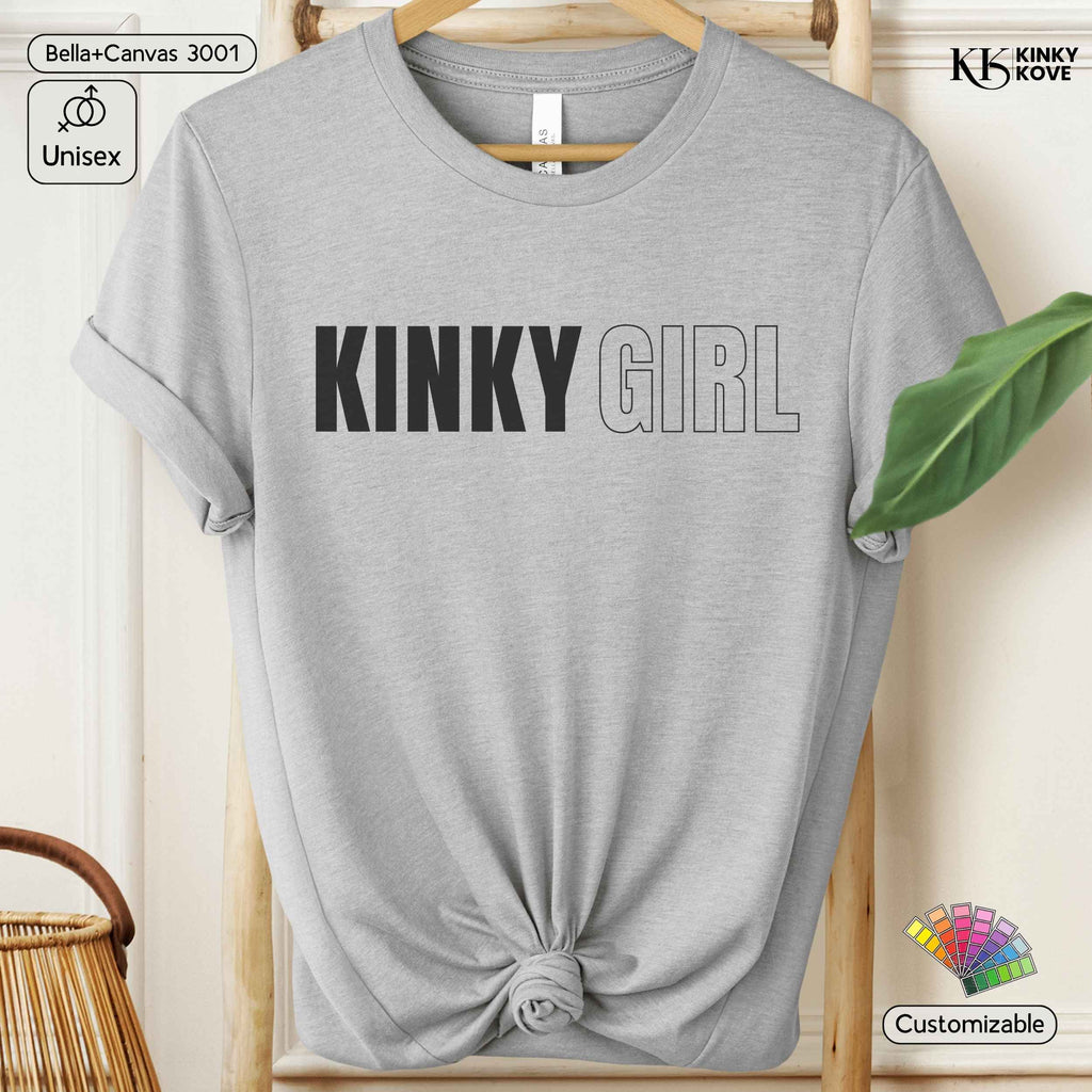 A light heather gray short-sleeve t-shirt with 'KINKY GIRL' printed across the chest, styled on a wooden hanger with a knotted hem. The image includes decorative elements like a green leaf, a wicker basket, and text badges indicating 'Bella+Canvas 3001', 'Unisex', 'KINKY KOVE', and 'Customizable'.