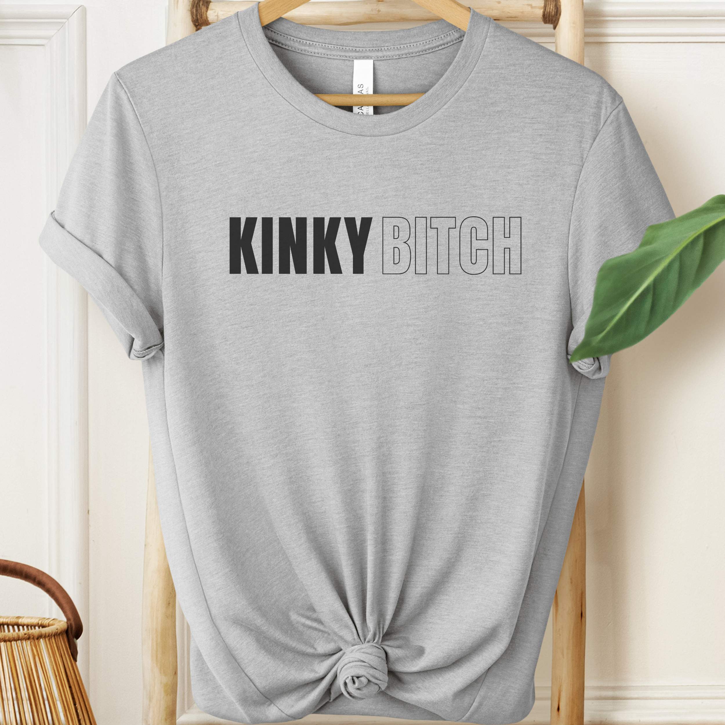 Gray t-shirt with 'KINKY BITCH' text on a hanger against a neutral background