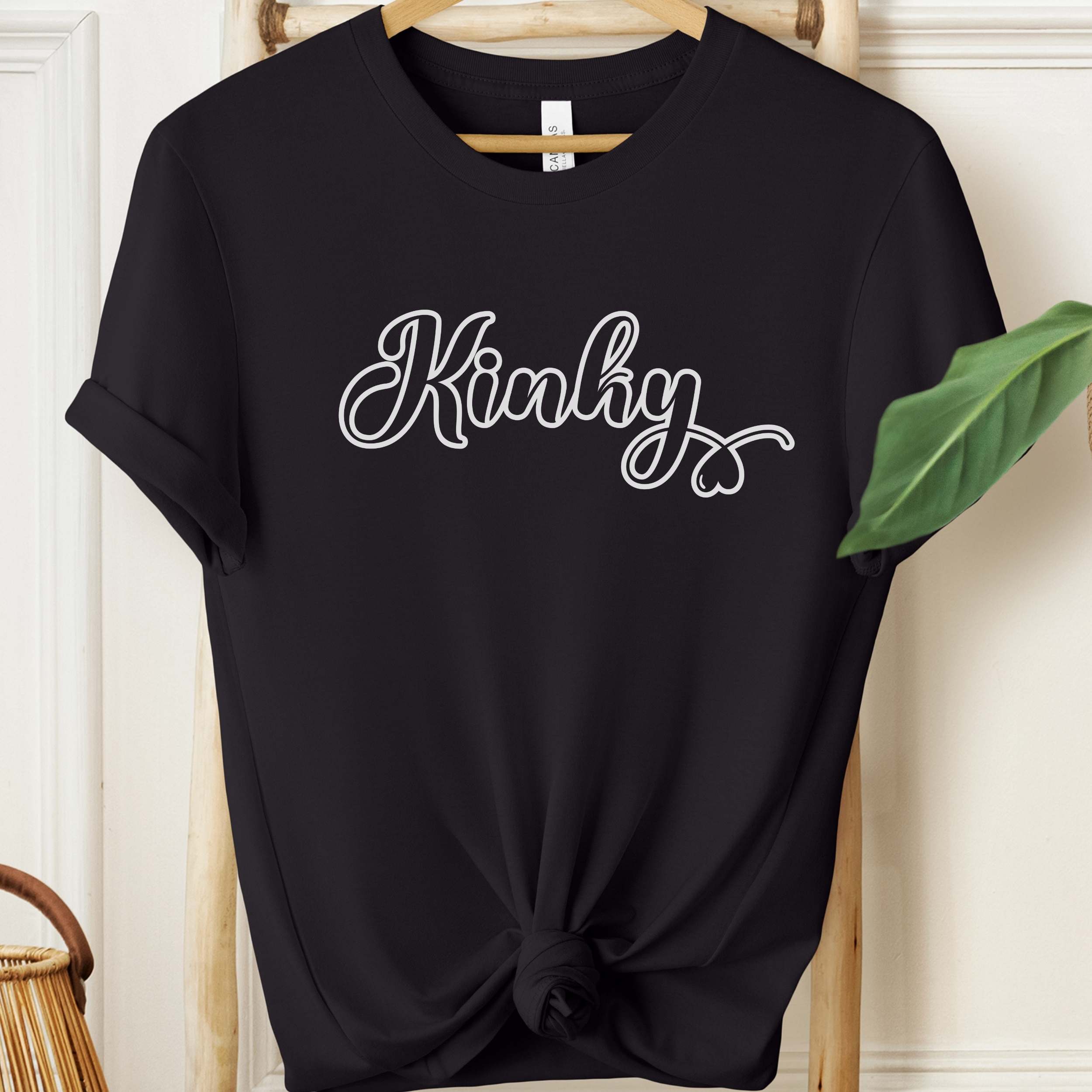 Black t-shirt with 'Kinky' text on a hanger against a white wall.